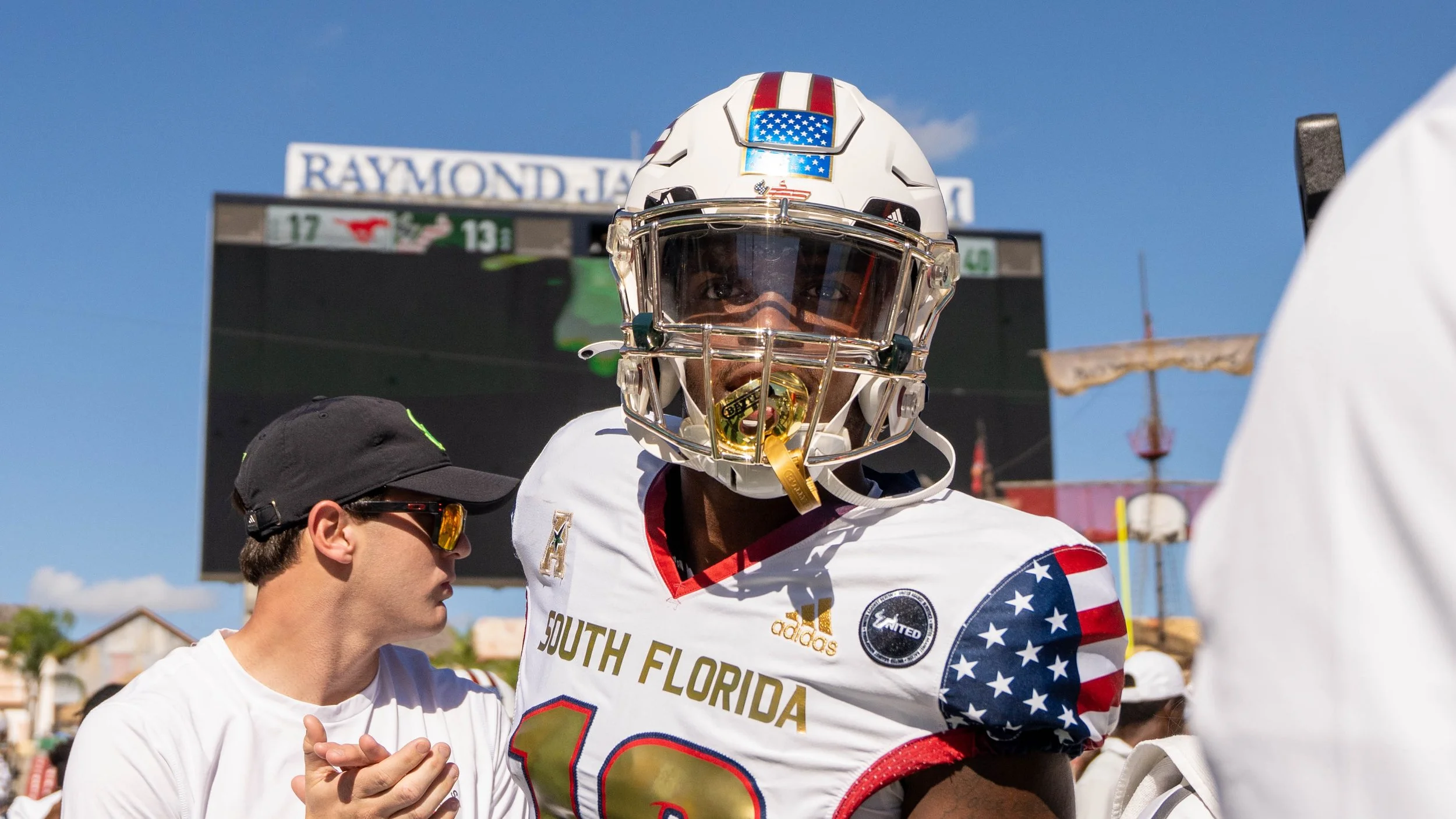 An American football player in a white uniform with red, blue, and gold accents, wearing a helmet with an American flag design, interacts with a person wearing sunglasses and a black baseball cap at a football game on a sunny day. There is a scoreboa