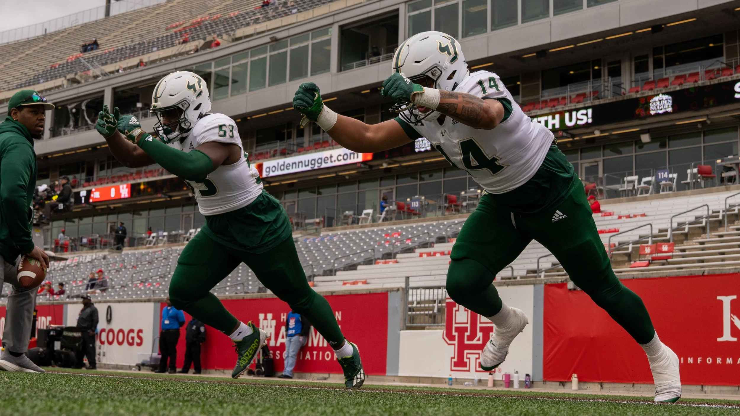 Two football players from the University of Houston team practicing on the field with a coach or trainer nearby, in a stadium with empty seating and a large digital scoreboard in the background.