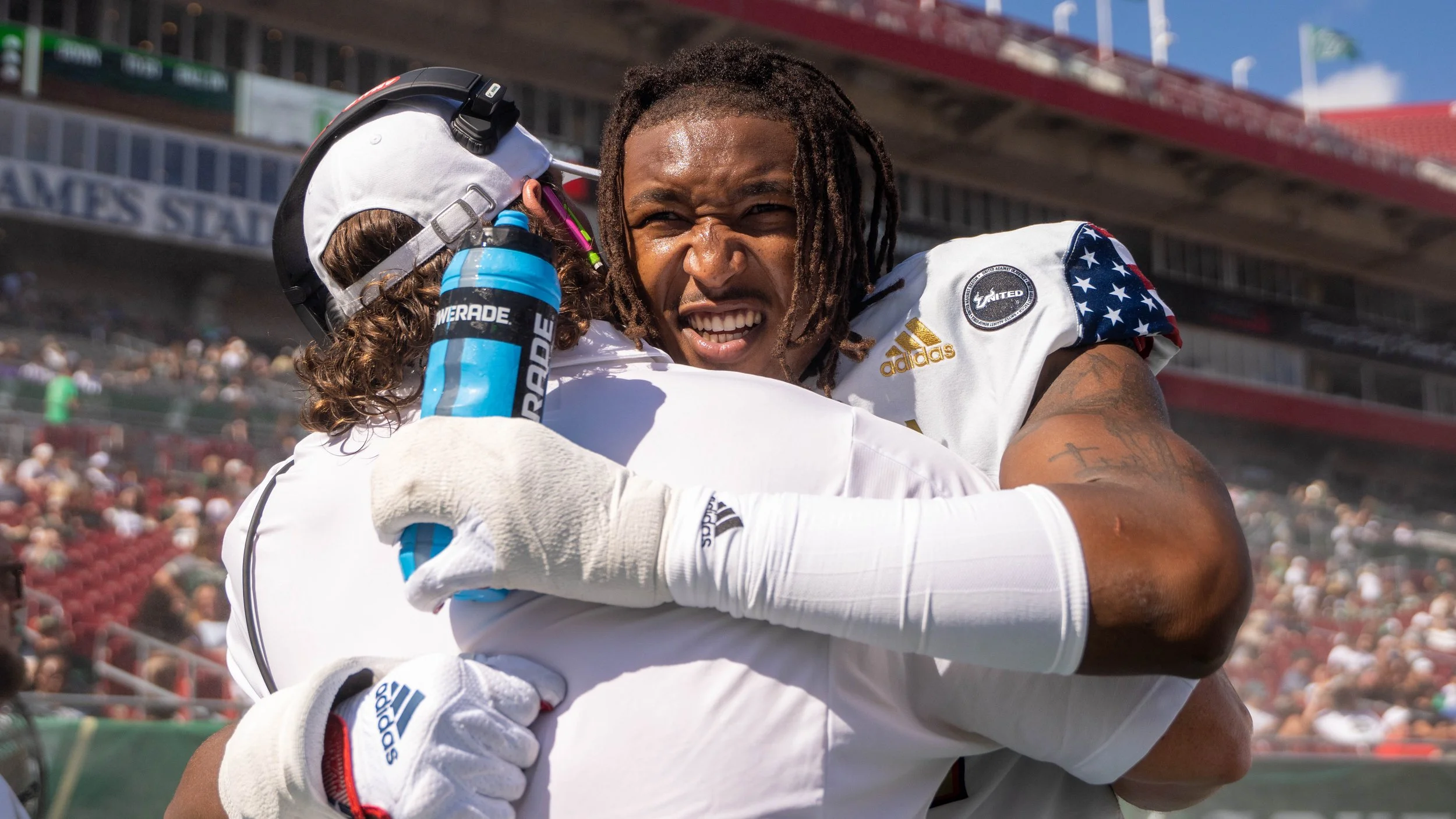 Two football players hugging on the field, one smiling with dreadlocks, the other wearing a helmet with a water bottle, in a stadium with spectators in the background.