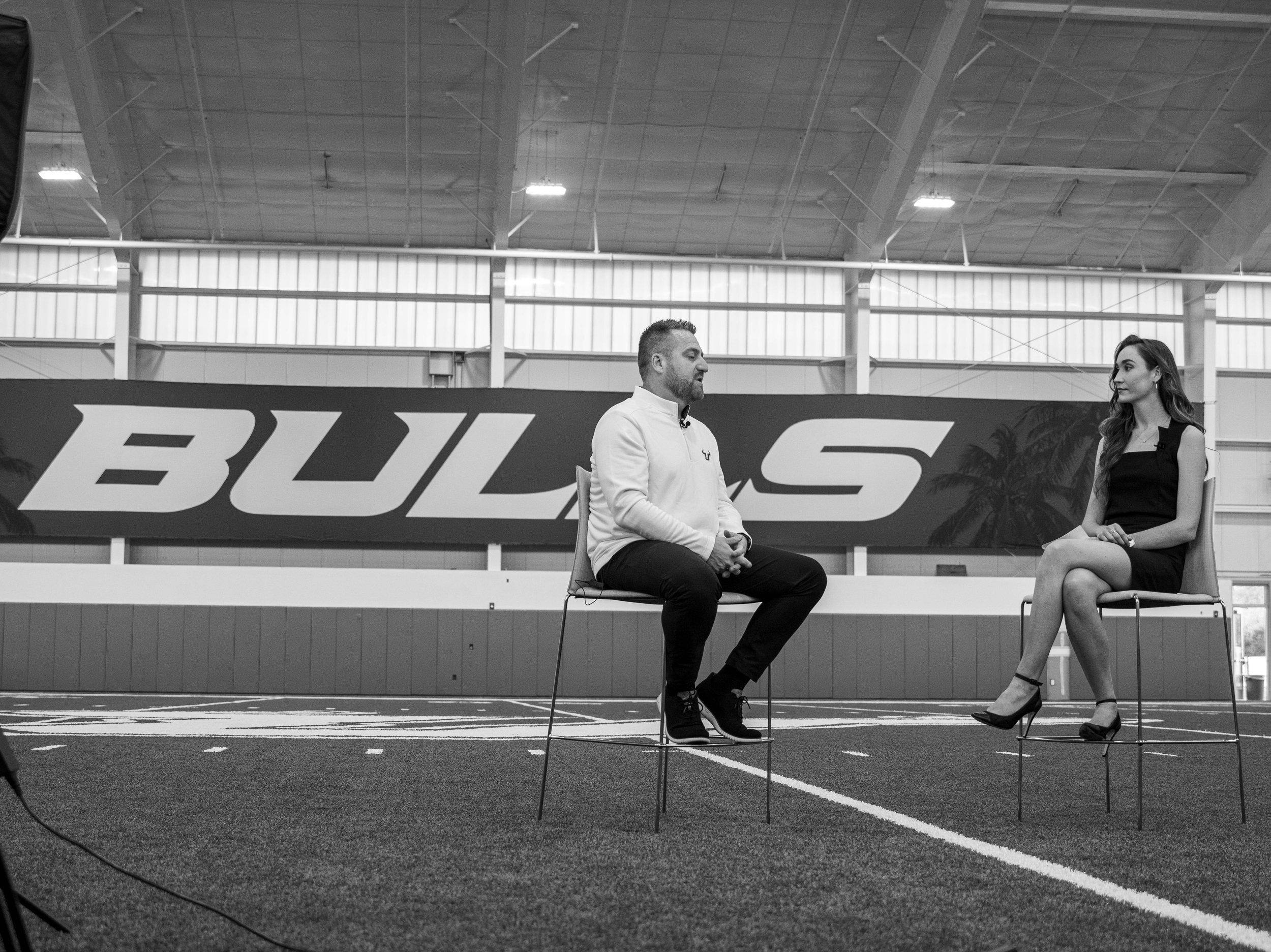 Two people sitting on chairs and having a conversation inside a sports arena or indoor athletic facility, with a large 'BUL' sign with palm tree graphics in the background.