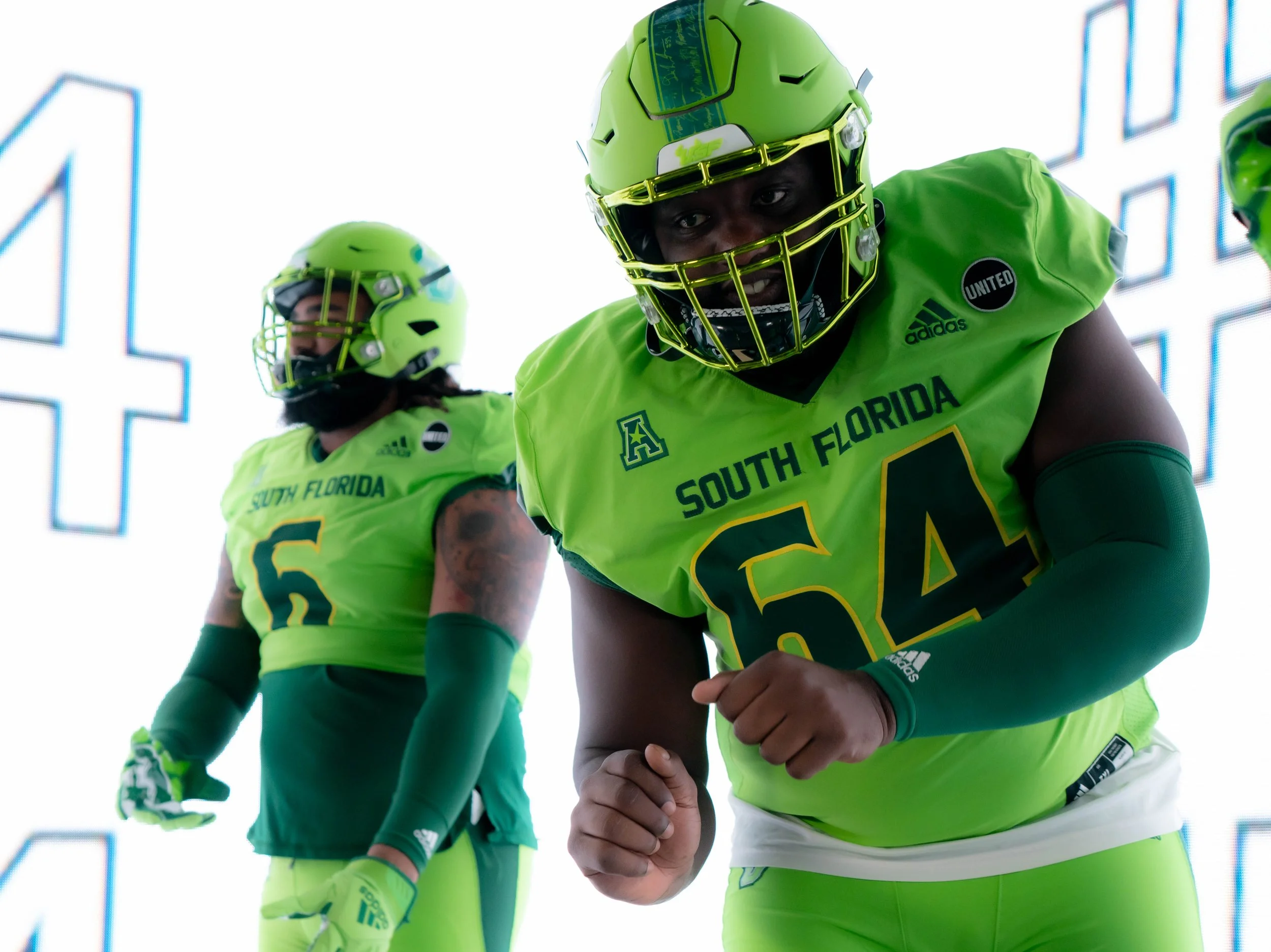 Two football players from South Florida in bright neon green uniforms and helmets, with one in the foreground showing a determined expression and clenched fists, and another in the background looking away.