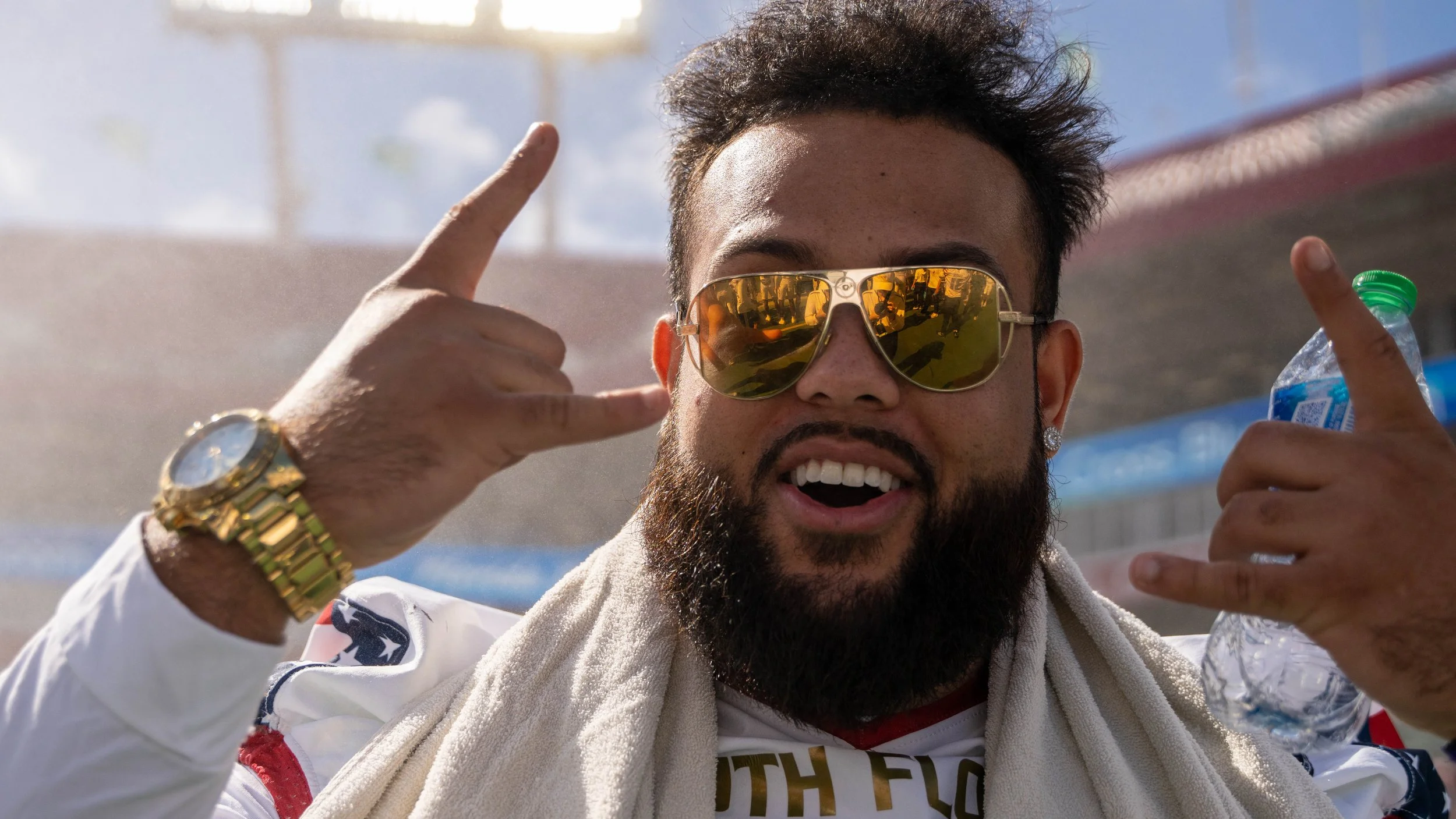 A man with a beard and curly hair wearing reflective sunglasses, a white sports jacket, and a gold watch, making a hand gesture with both hands near his face, holding a water bottle in his right hand outdoors.