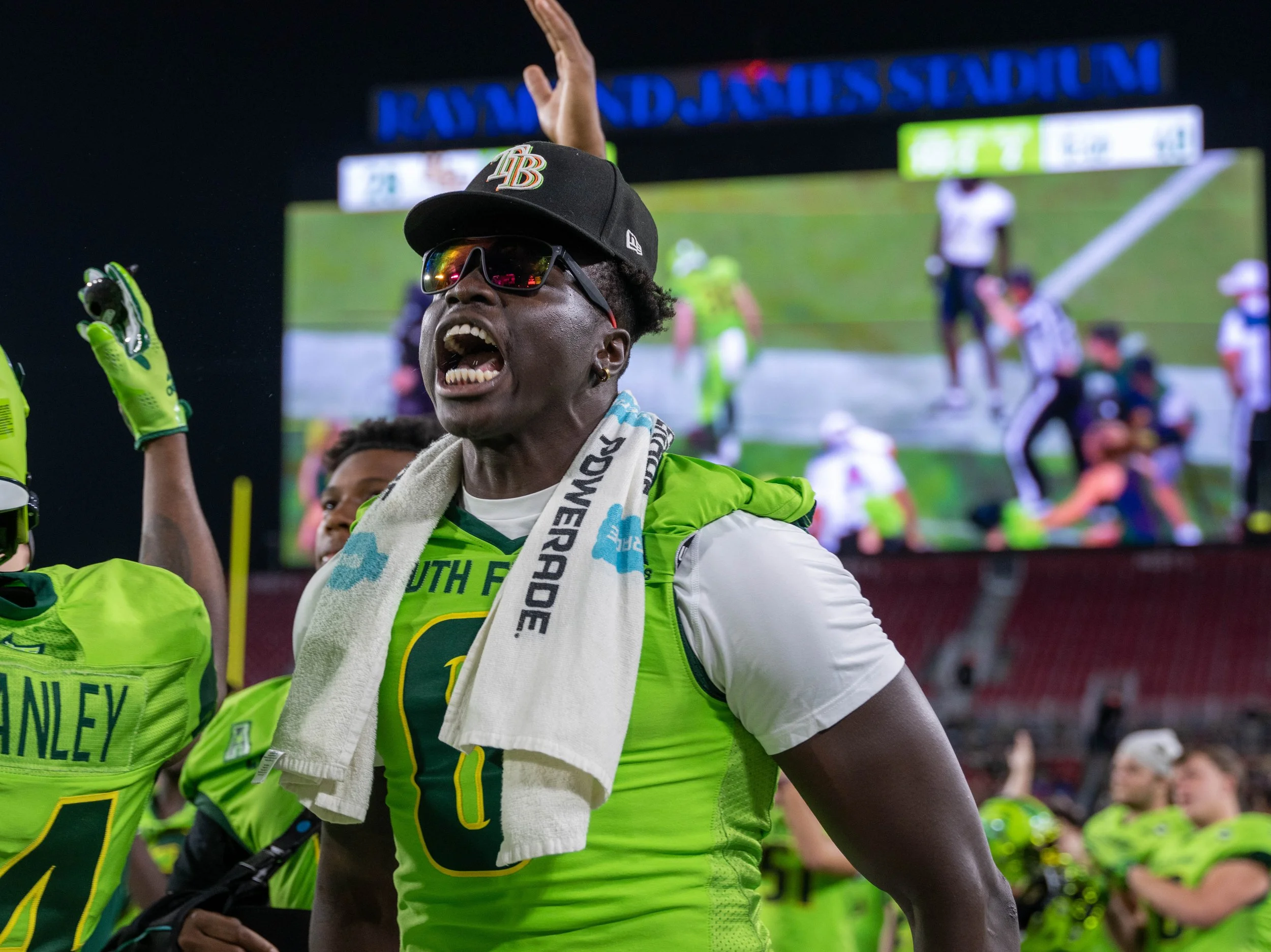 A football player in a neon green uniform celebrating on the field at Raymond James Stadium, with a towel around his neck, sunglasses, and a cap, during a night game.