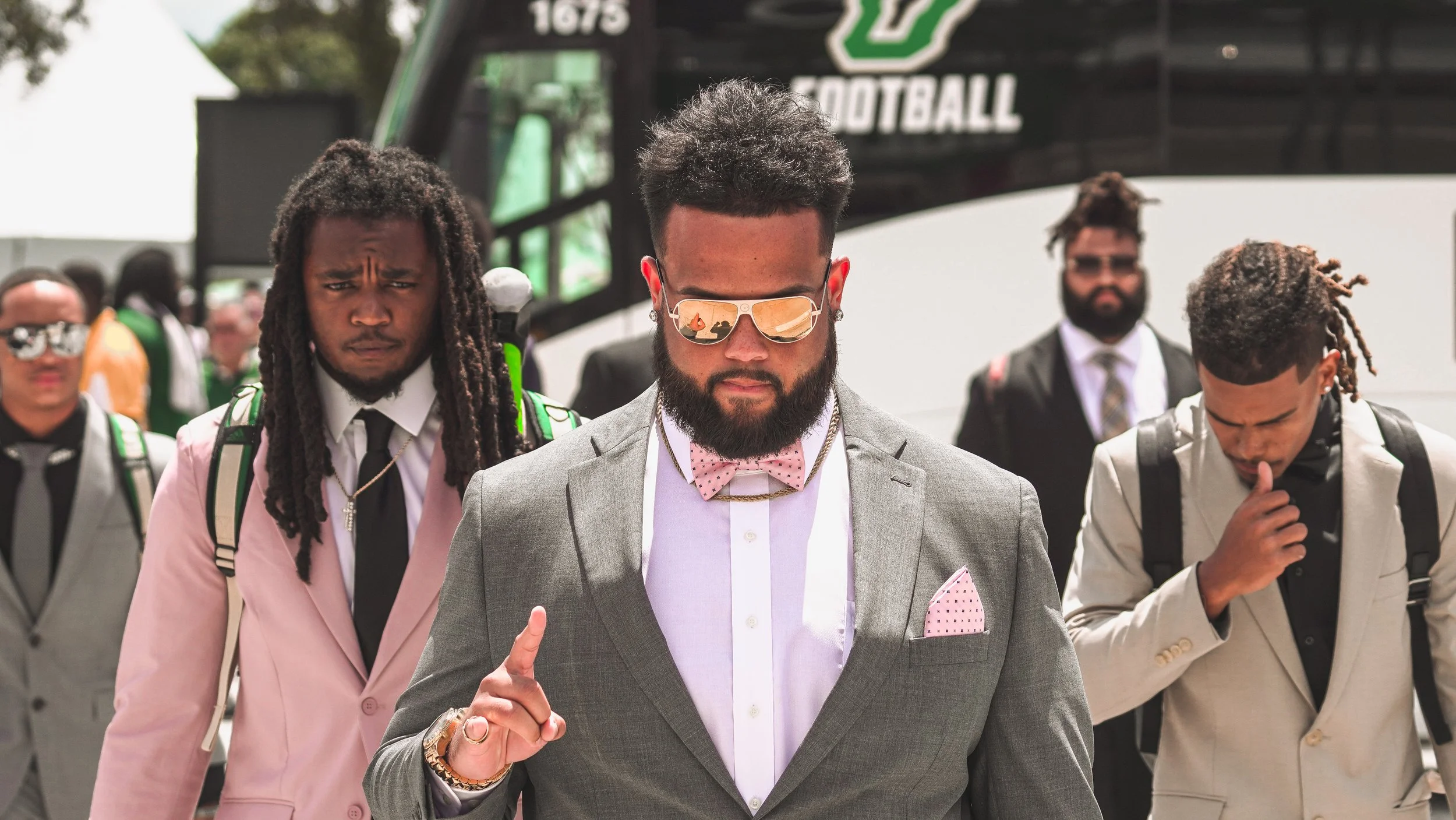 A group of five stylish men walking outdoors, with a bus in the background. The man in front wears sunglasses, a gray suit, a pink bow tie, and has a beard.