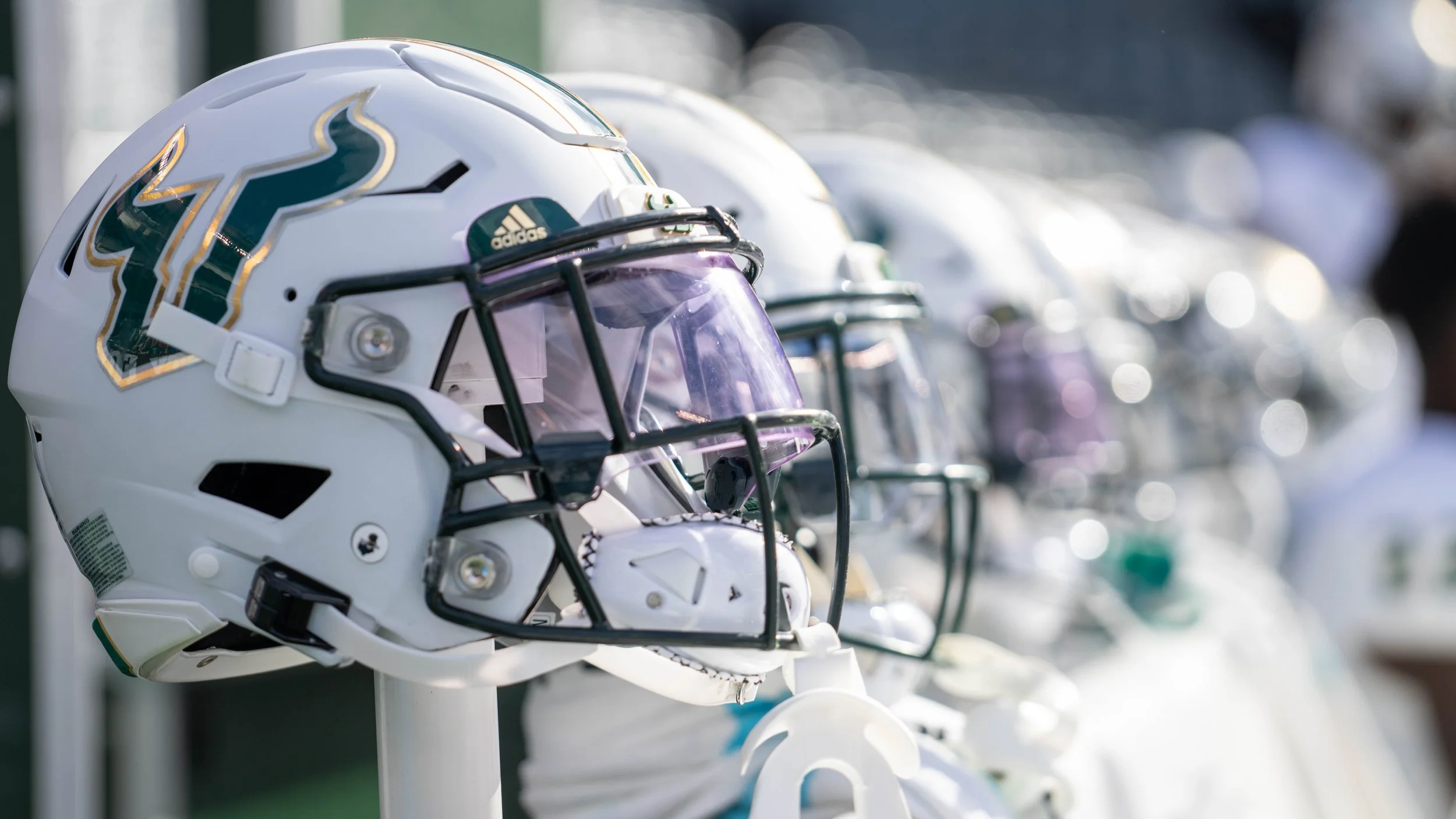 A close-up of a white football helmet with a gold and green logo, placed on display among other helmets in the background.