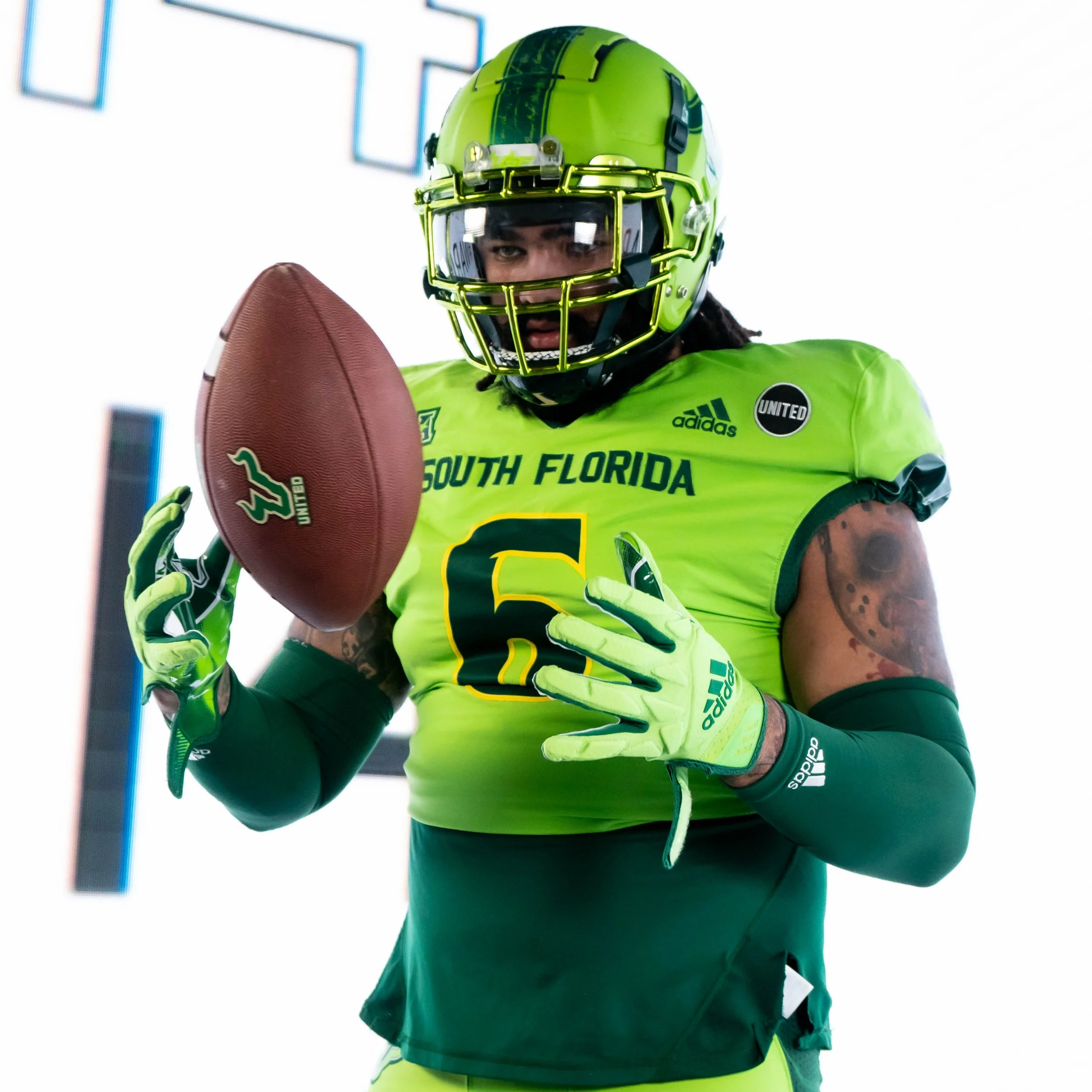A football player in bright green uniform holding a football, with a helmet and gloves, looking down at the ball.