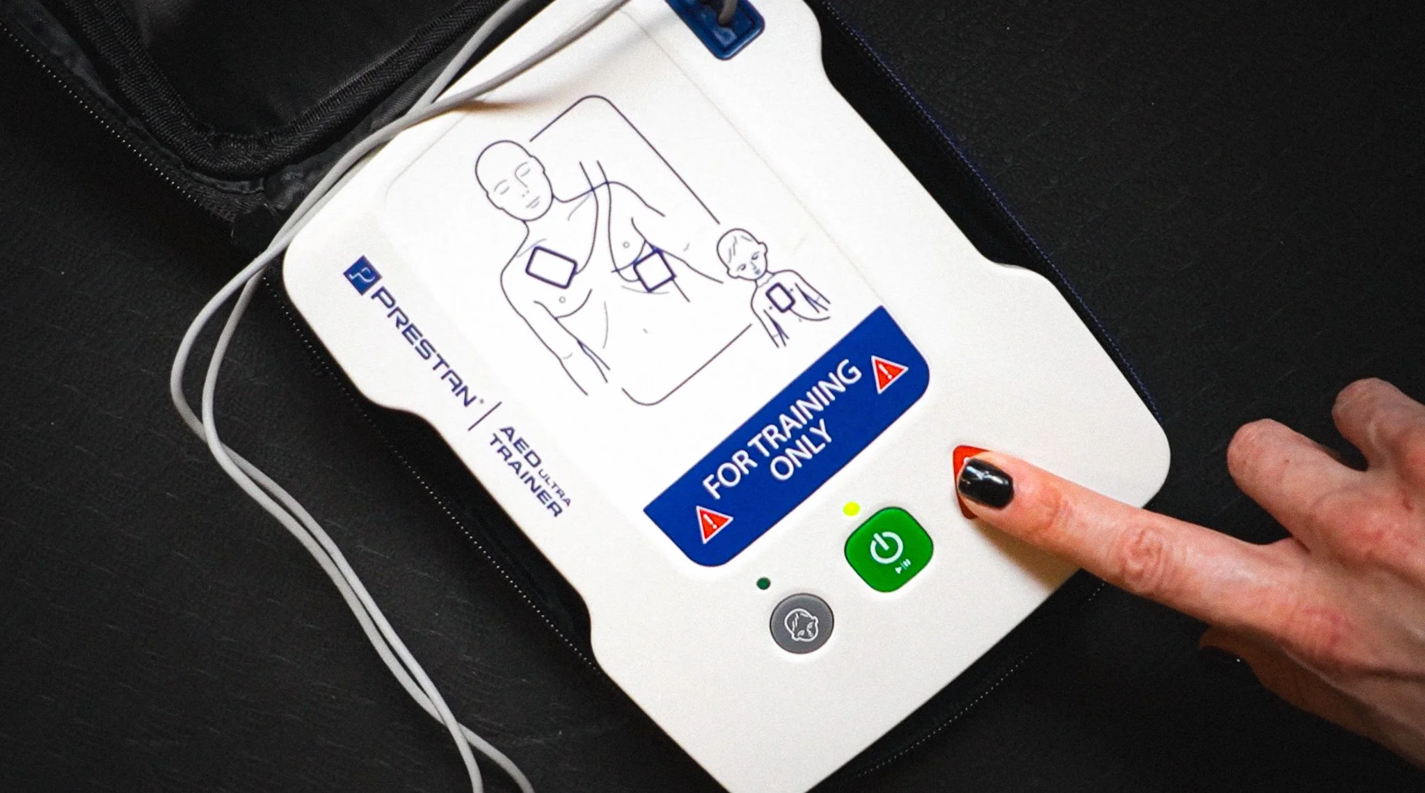 Automated external defibrillator (AED) device with a graphic illustrating its use, labeled for training purposes only, resting on a black surface, with a person's hand pressing the power button.