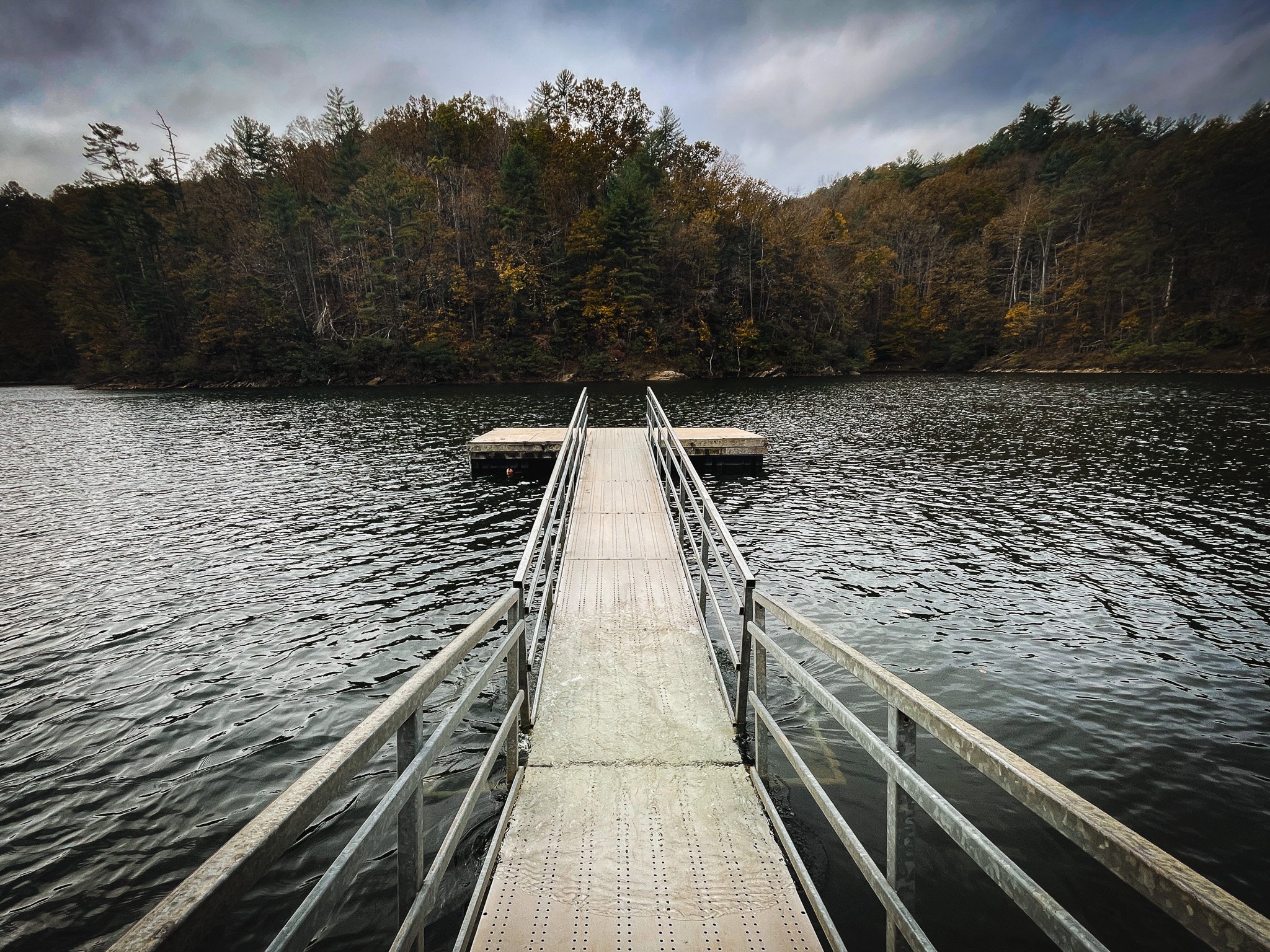 A metal dock extends into a body of water, with trees in fall colors on the opposite shore under a cloudy sky.