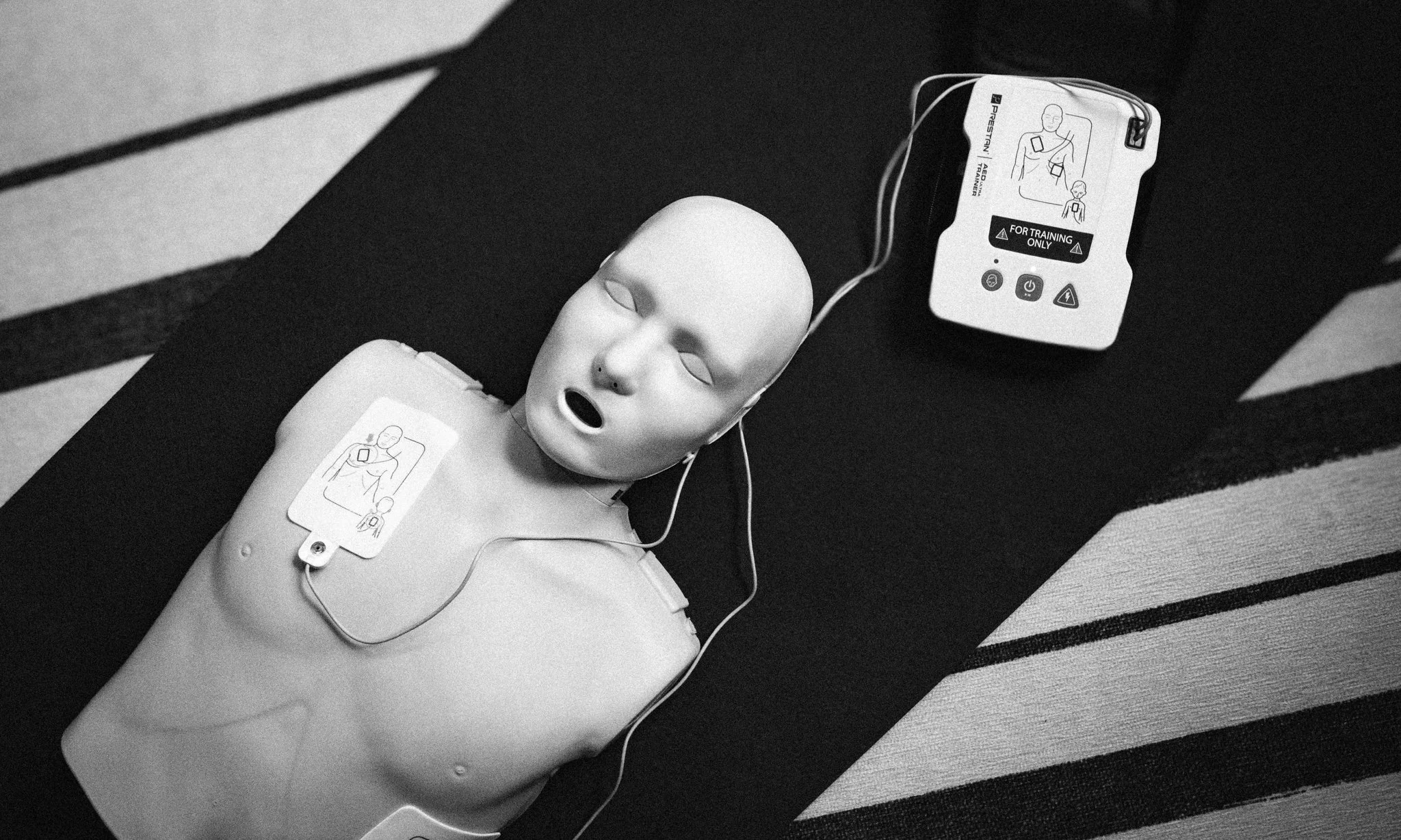 A medical training mannequin with a mask and wires connected to a defibrillator for simulation practice on a striped mat.