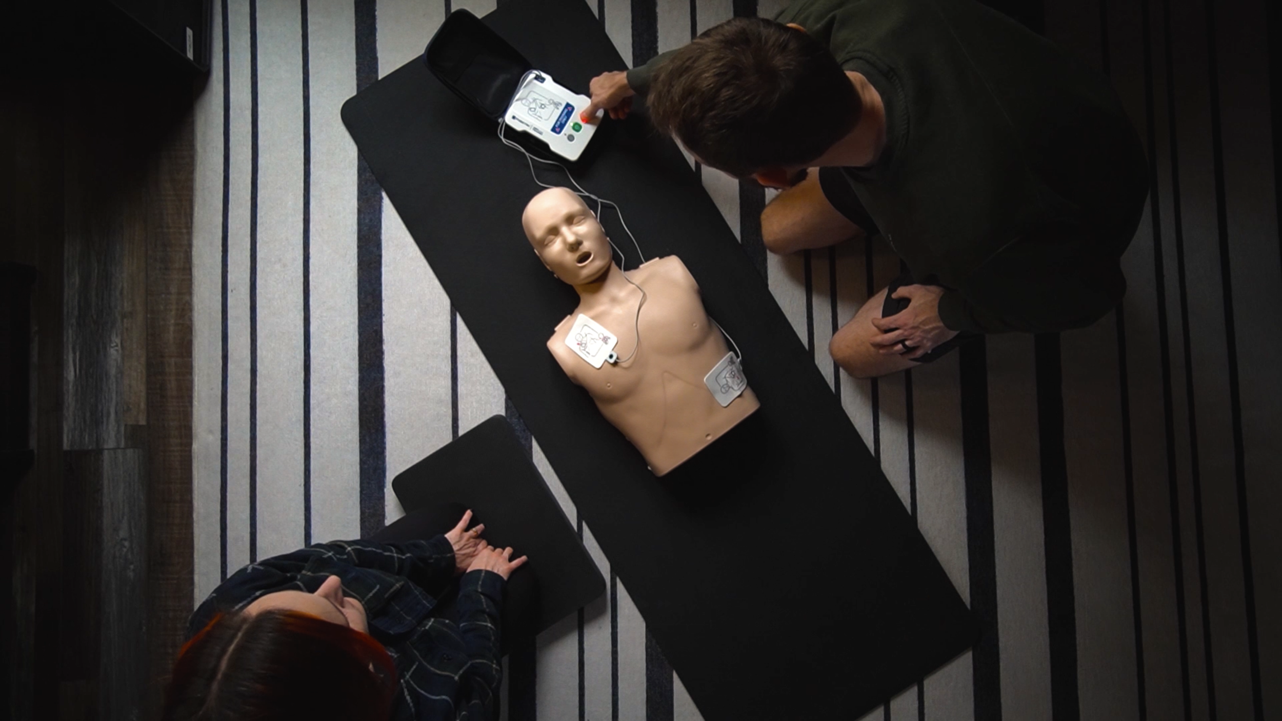 A man practicing CPR on a medical training dummy lying on a table, with a woman and another man watching.