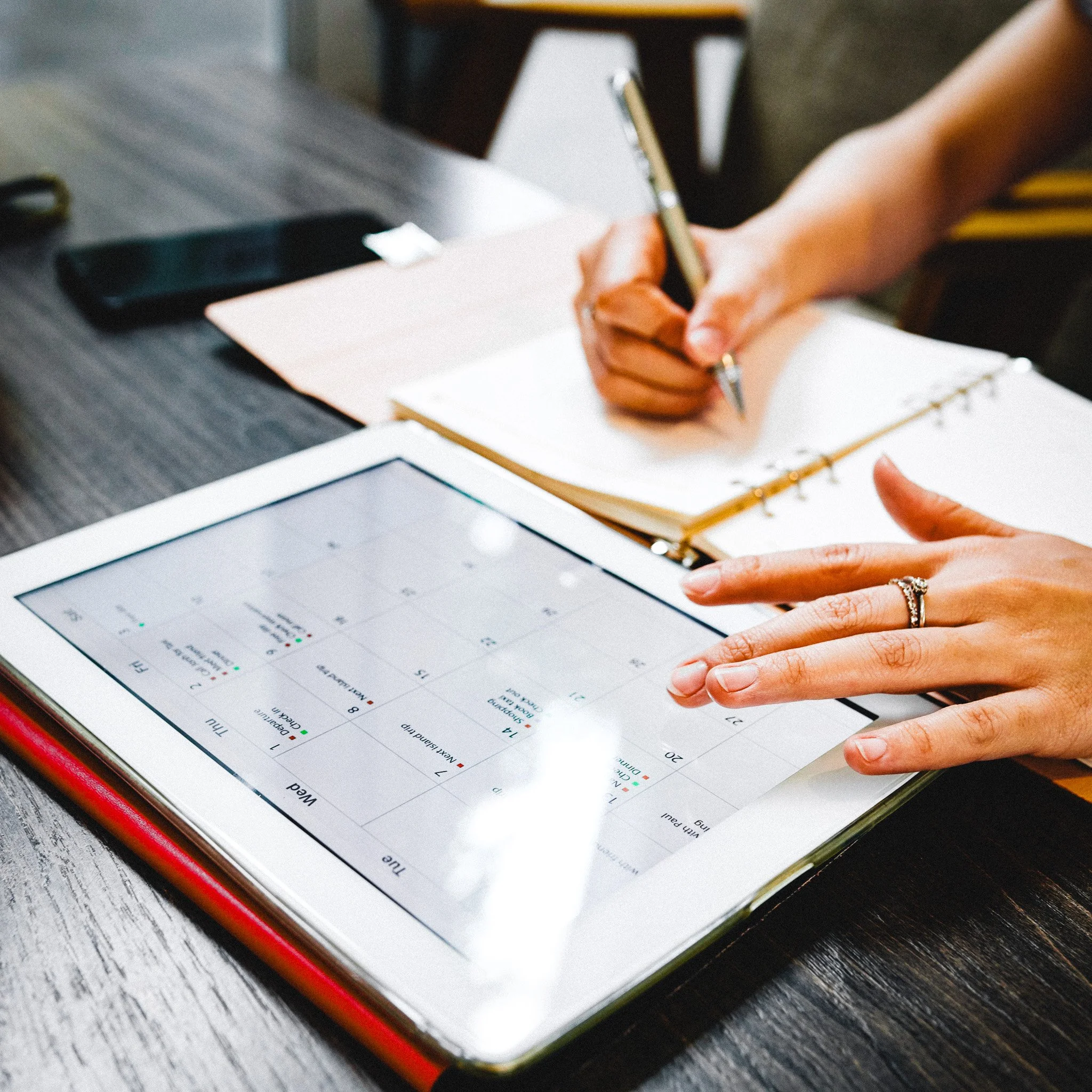 Person using a tablet showing a calendar with dates and tasks, while writing in a notebook with a pen on a dark wooden table.
