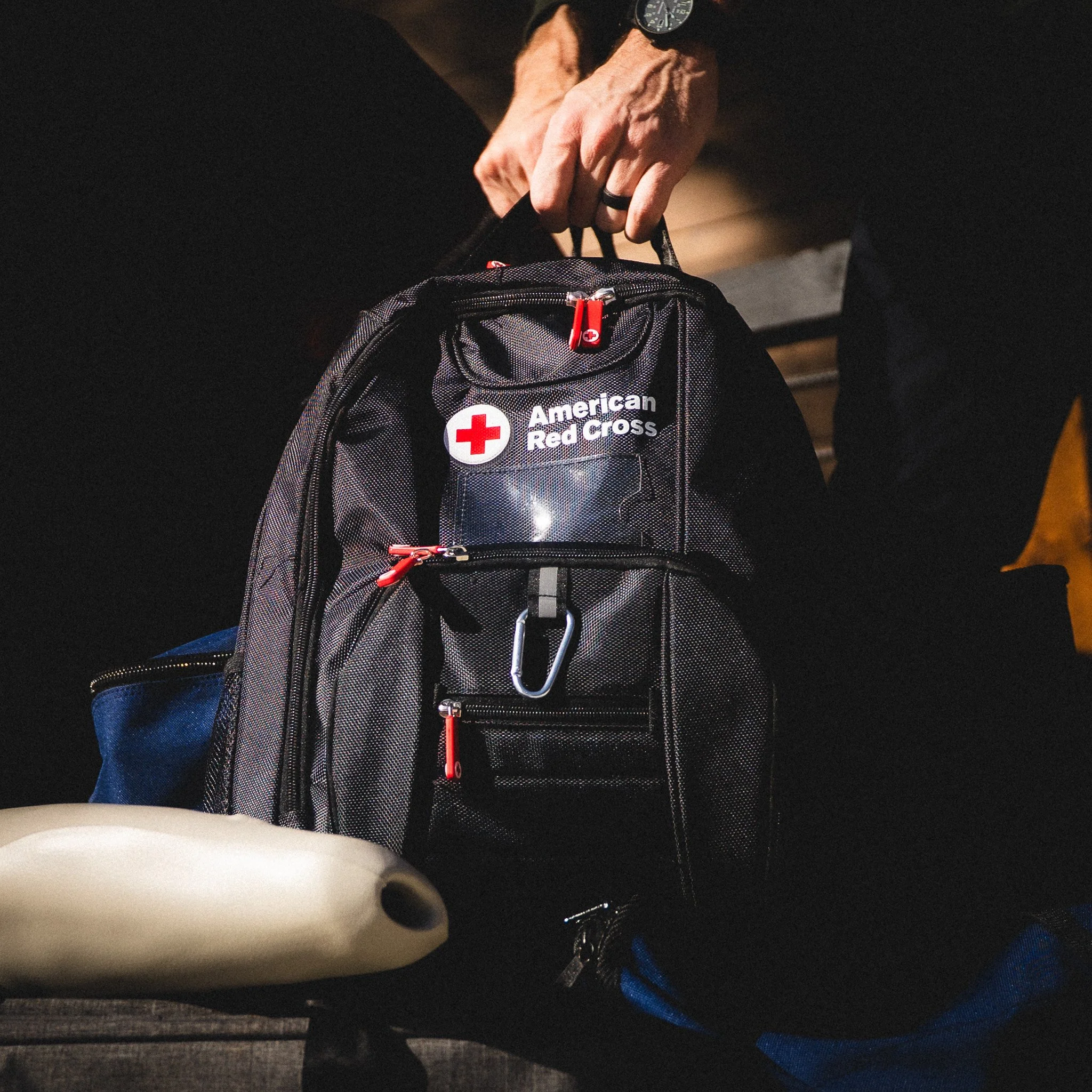A person holding a black backpack with an American Red Cross logo, with various compartments, zippers, a carabiner, and a clear plastic pocket. The person is wearing a watch and a ring, and part of their arm is visible.