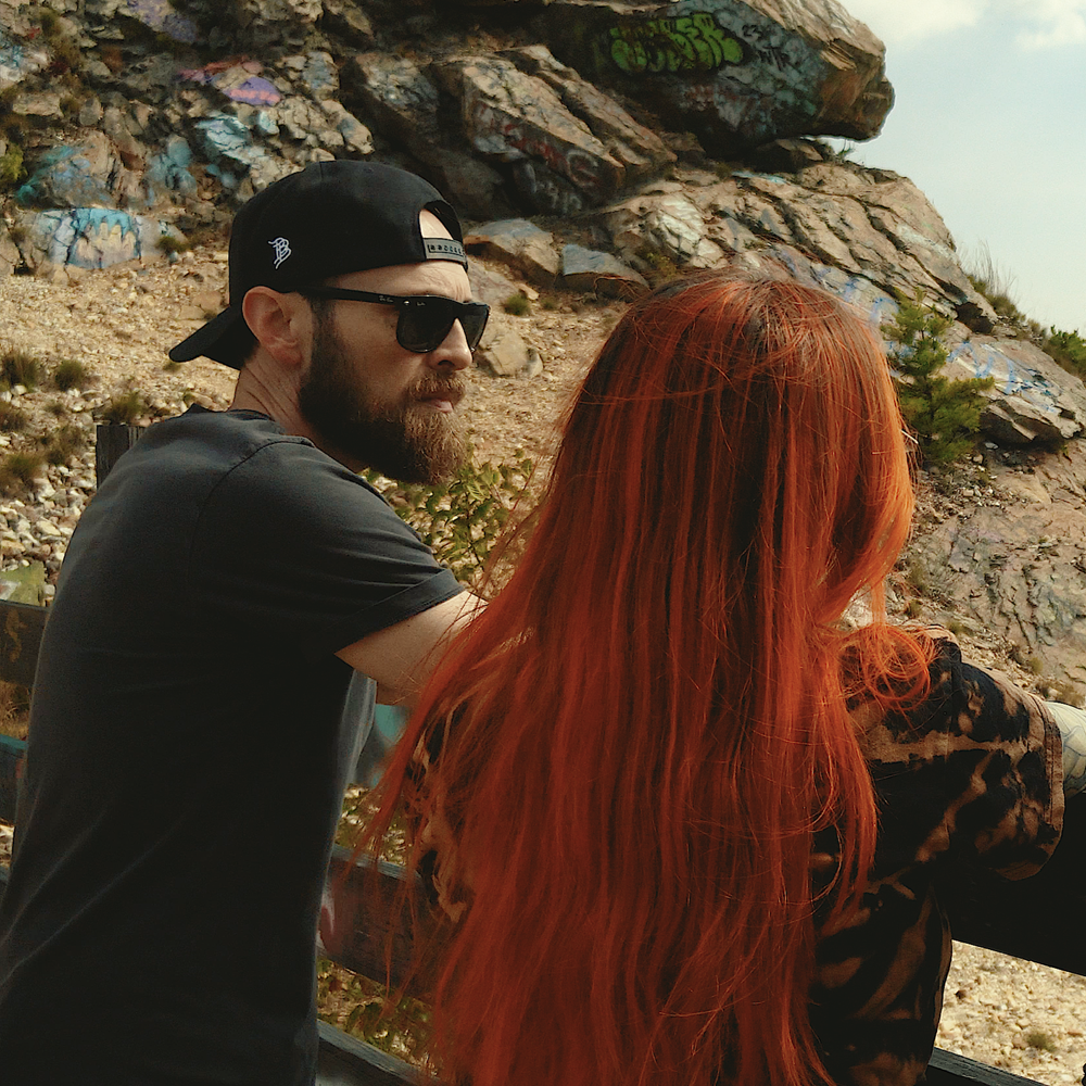 A man and a woman with long red hair are sitting outdoors near a rocky hillside with graffiti, looking towards the distance.