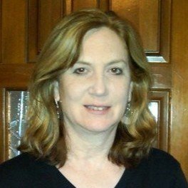 A woman with shoulder-length wavy blonde hair wearing a black top, standing indoors in front of wooden cabinets.