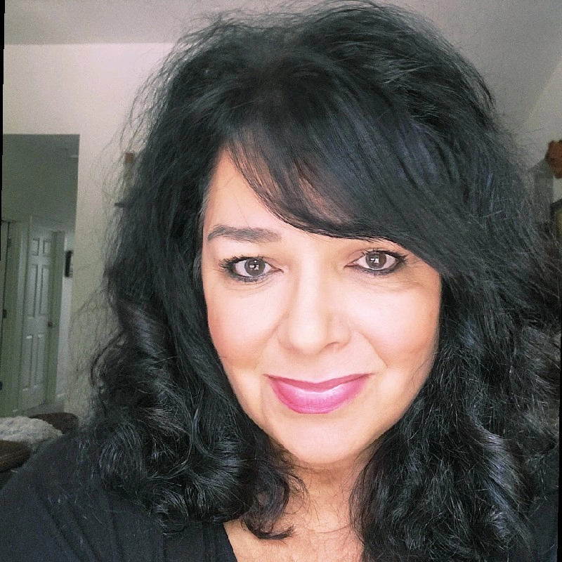 A woman with wavy black hair and blue eyes smiling at the camera, indoors with a background of a room with light-colored walls and an open door.