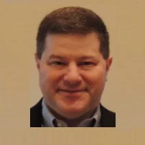 Portrait of a man with short brown hair, smiling, wearing a dark blazer and light shirt, against a plain background.