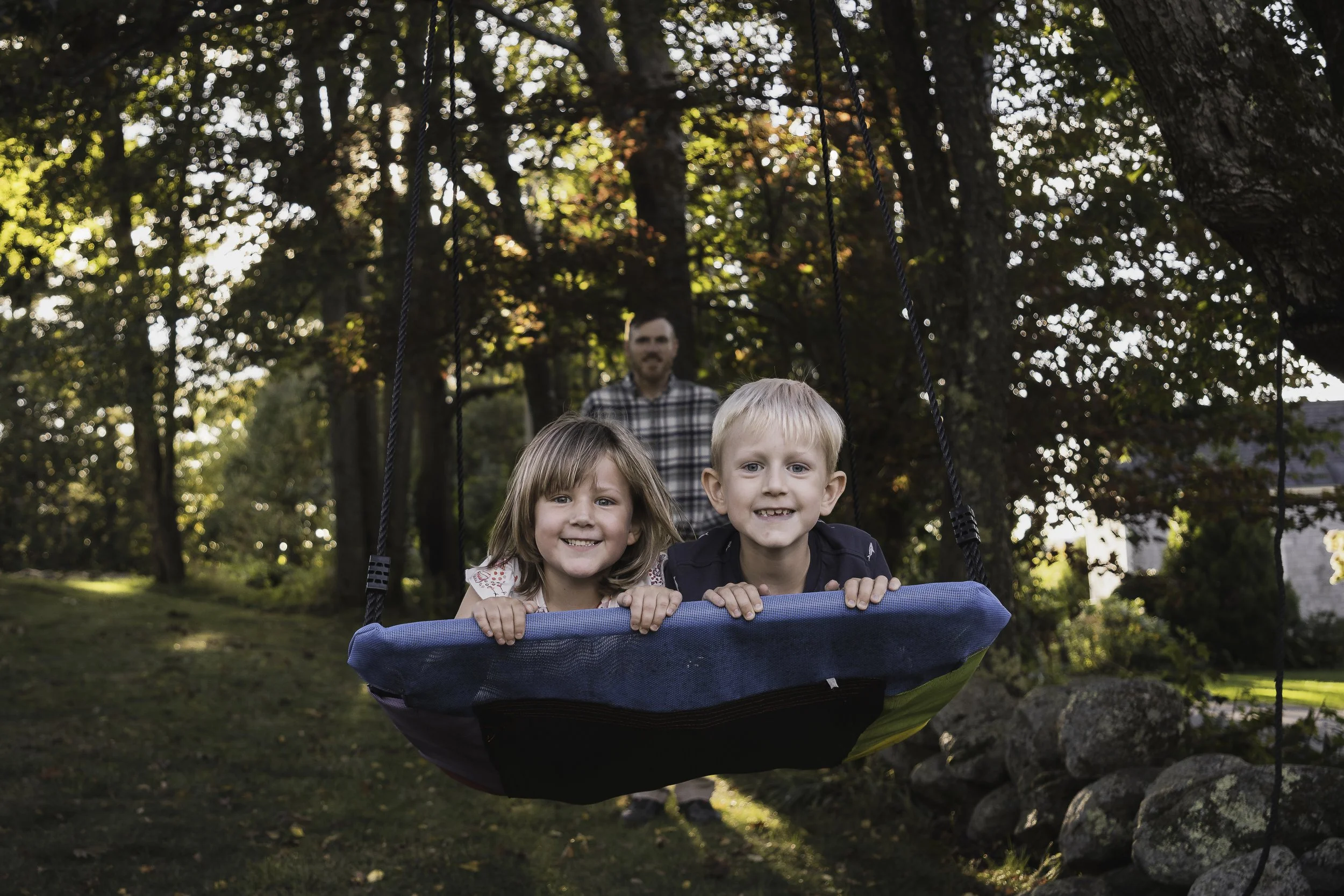 Candid outdoor family photography session in Central Massachusetts featuring siblings smiling on a swing at golden hour.
