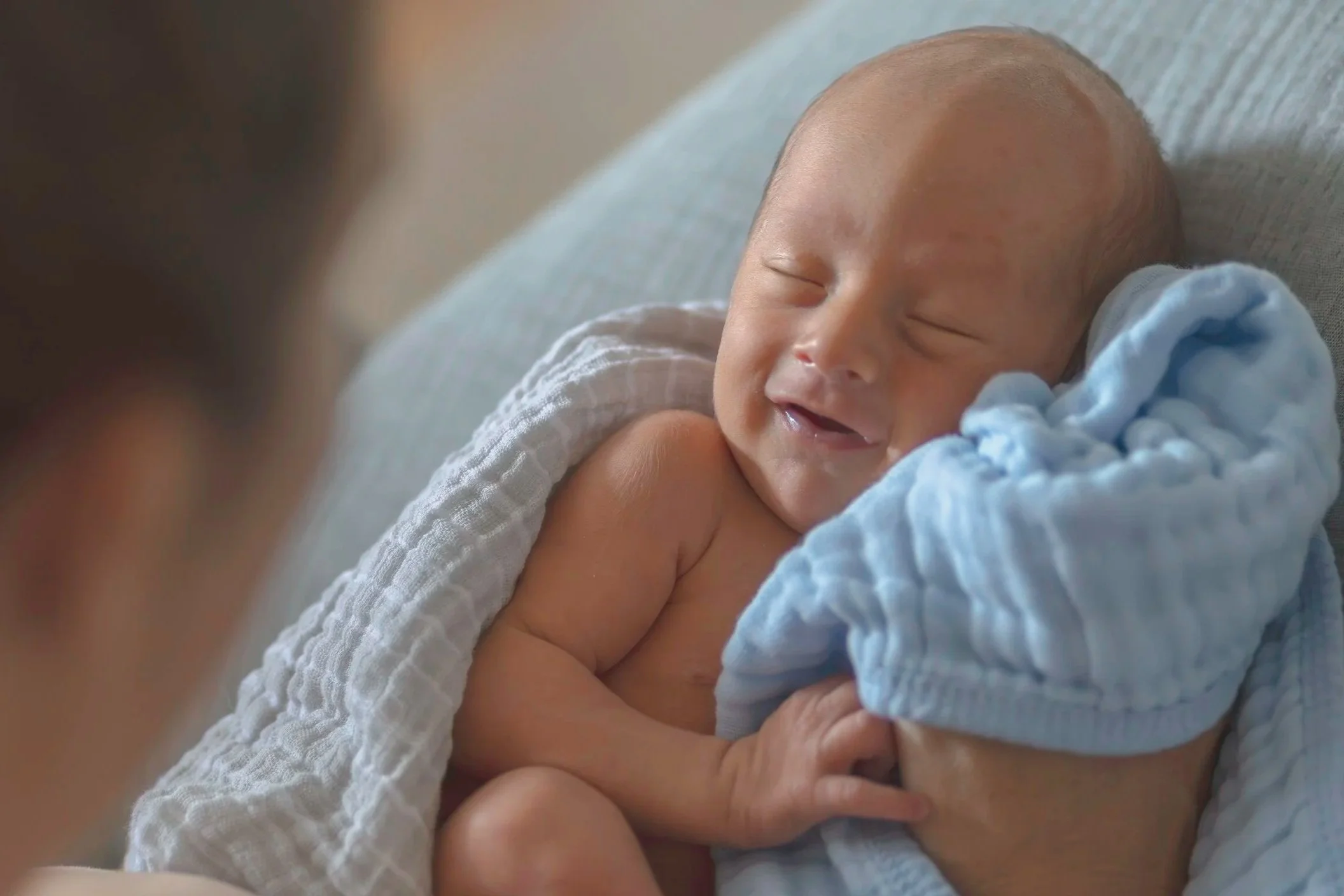 During a lifestyle newborn photo shoot near Westminster Massachusetts a smiling newborn baby with a dark head of hair, wrapped in a light blue blanket, lying on in mom's arms.  