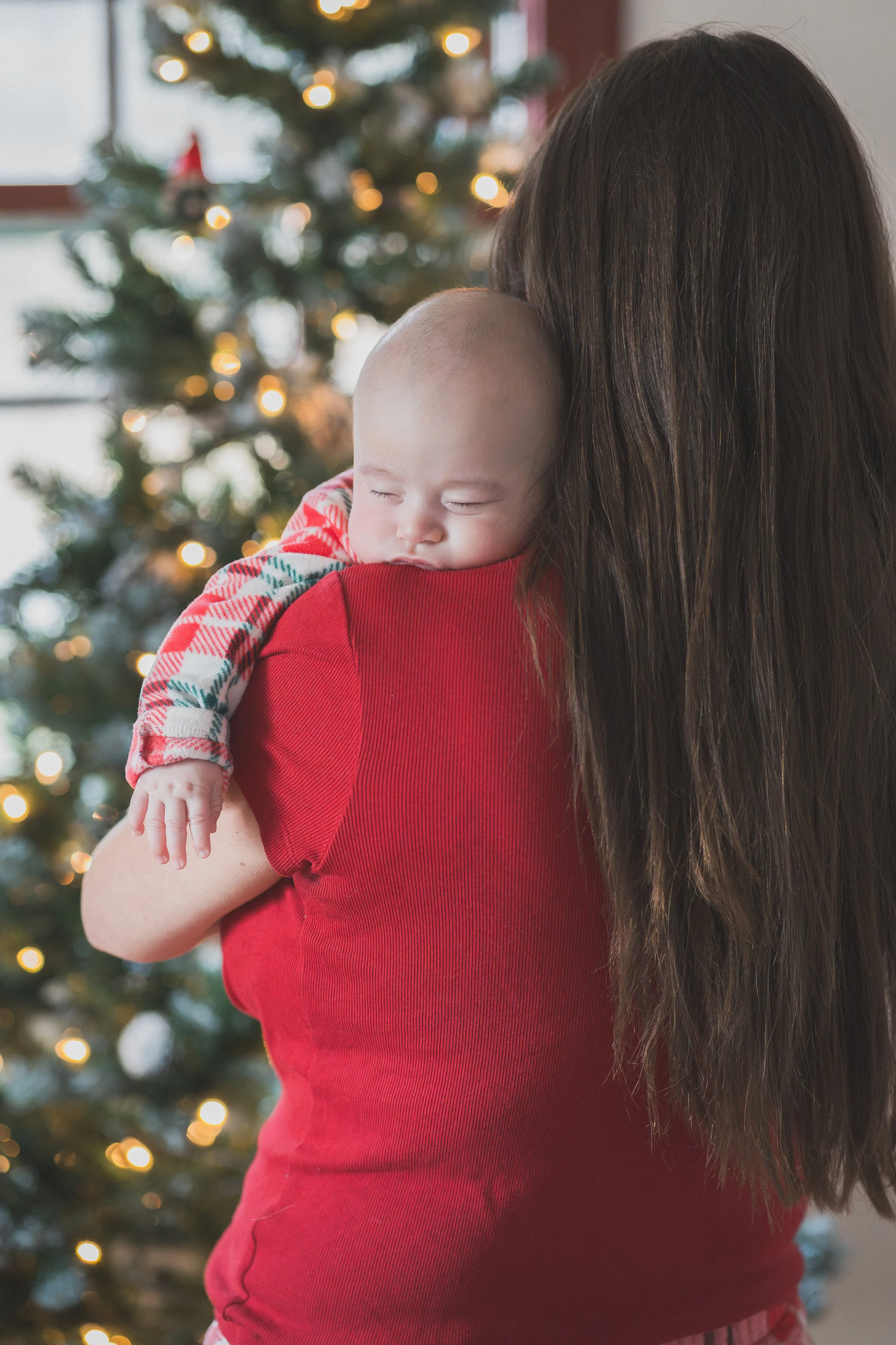 Sleeping baby in red plaid pajamas resting on mother’s shoulder beside a decorated Christmas tree.