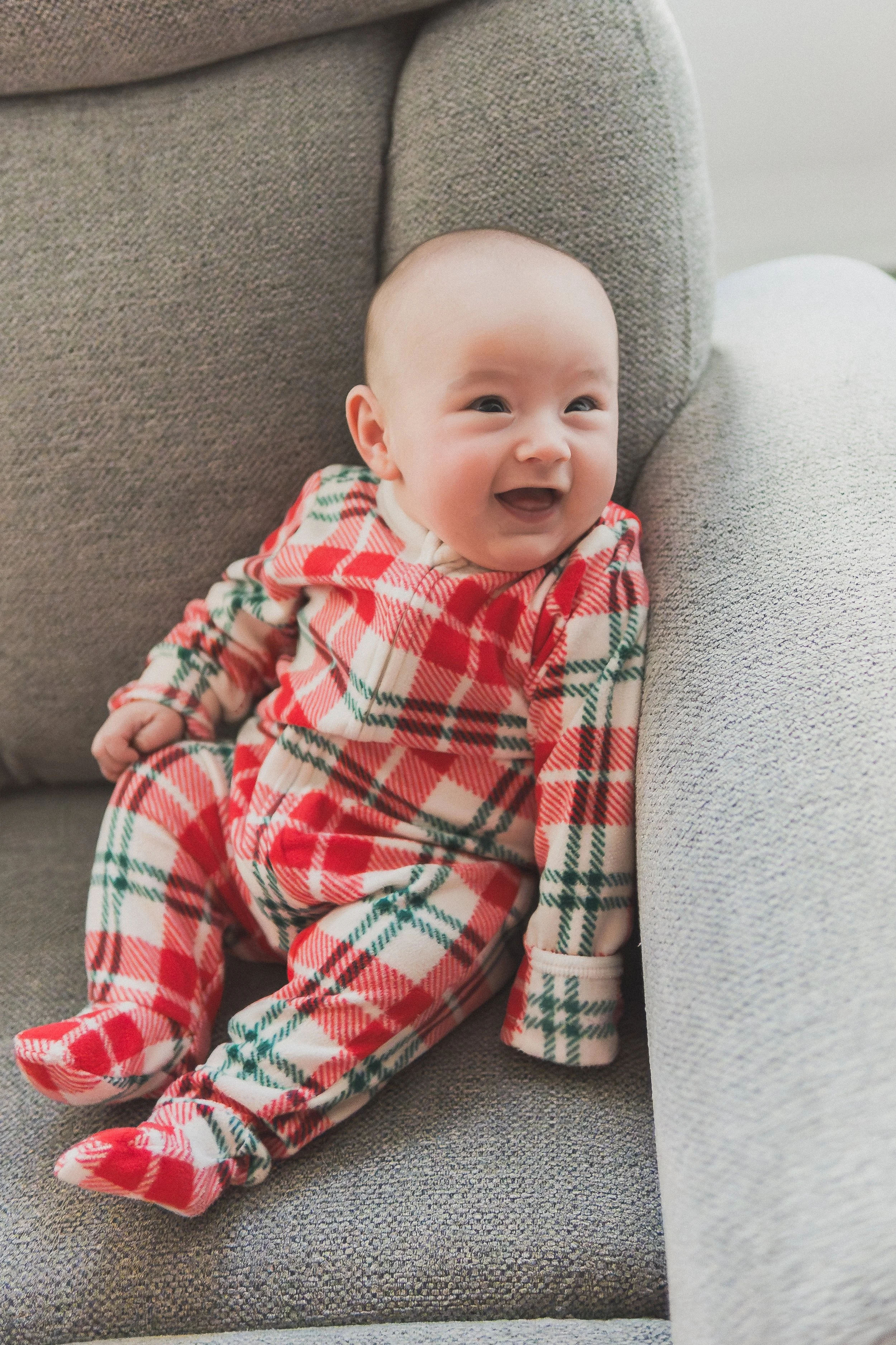 Smiling baby in red plaid pajamas sitting upright in a chair during a cozy holiday lifestyle session.