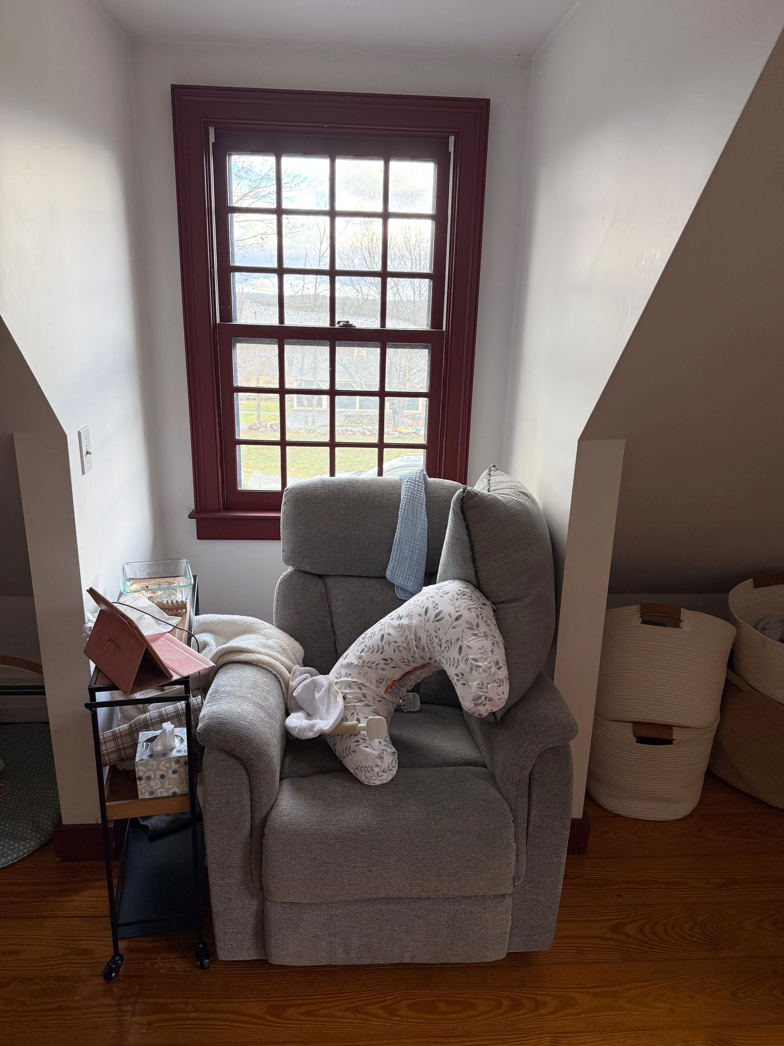 A nursery reading corner before cleanup, showing a gray armchair, baby pillow, basket, and natural window light — an example of a lived-in space before preparing for an in-home family photo session.