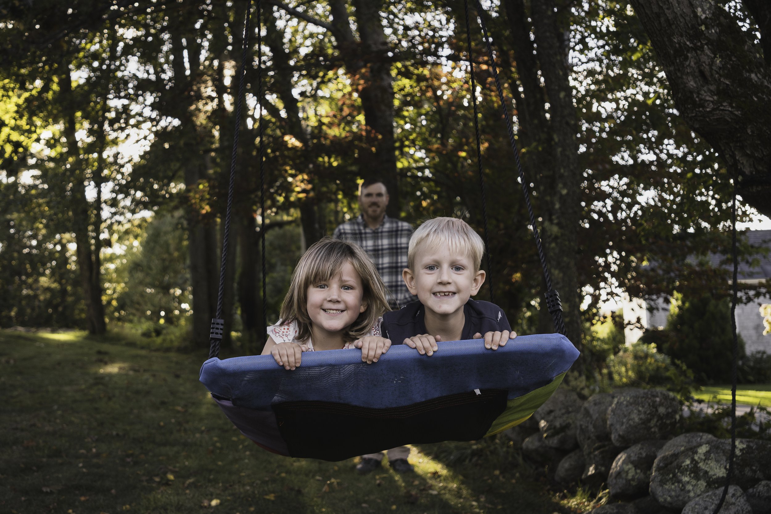 Young brother and sister smiling together on a swing during a golden-hour family photo session in Central Massachusetts, with their father in the background. Lifestyle family photography capturing joy and connection.