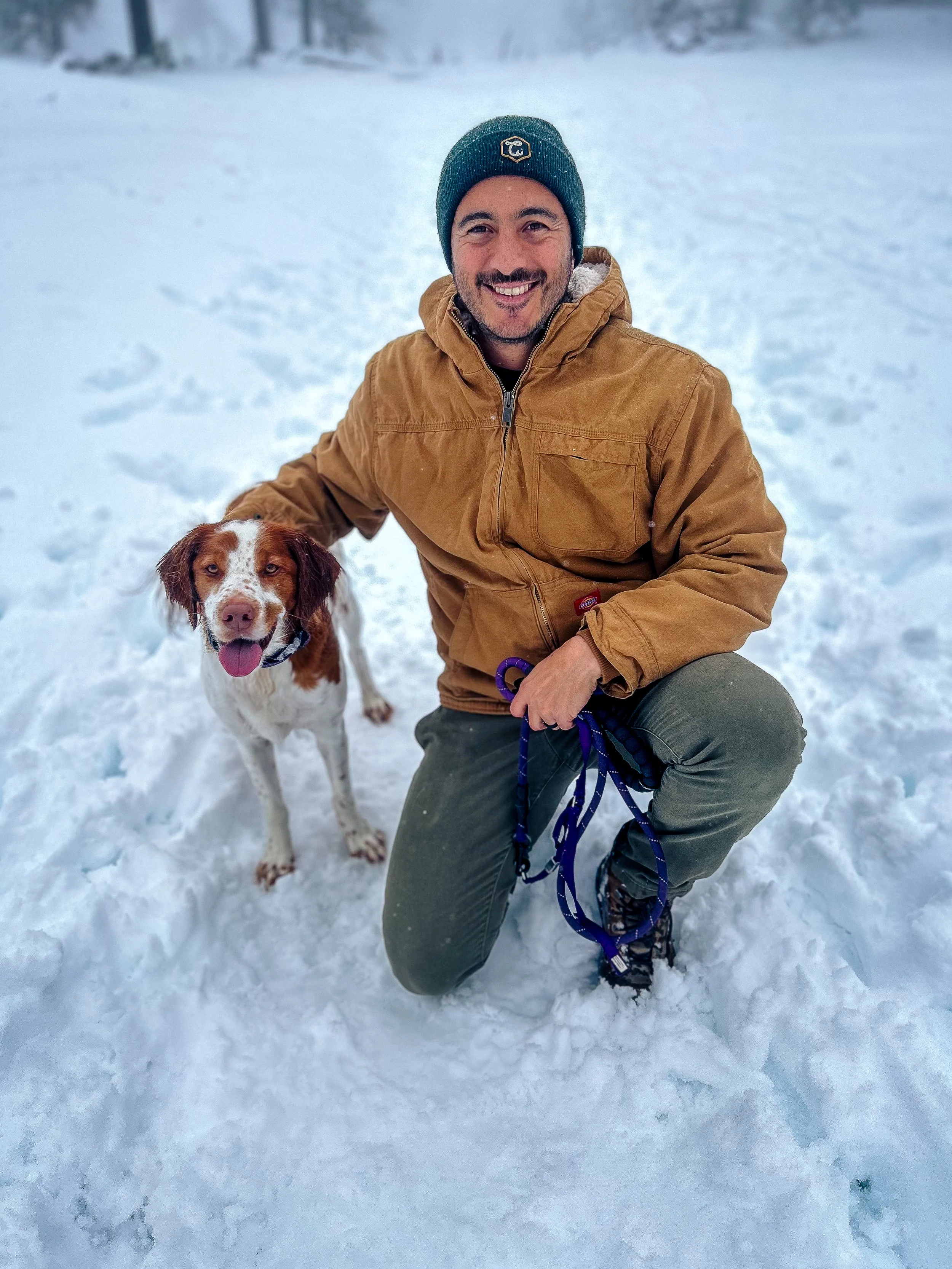 Your Central Mass family photographer kneeling in the snow with a brown and white dog, both smiling at the camera.