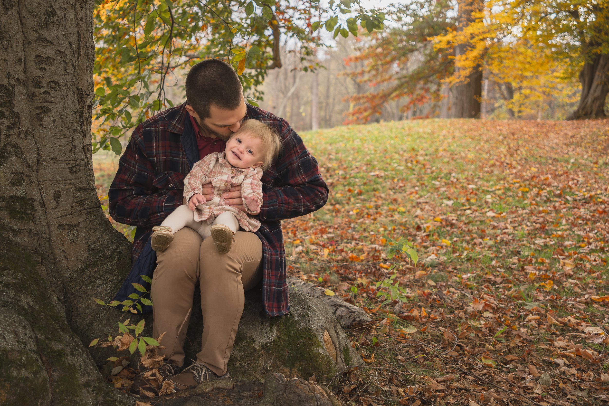 A father and daughter sitting on a large tree root in a park during autumn.  The girl is smiling and looking up. The background features fallen leaves and colorful trees with orange and yellow foliage common in Concord Massachusetts.