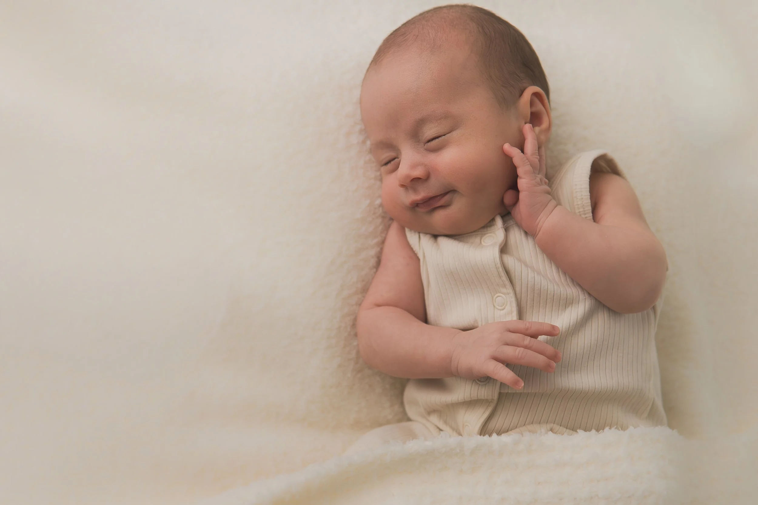 Smiling newborn lying on a soft light-colored blanket, wearing a sleeveless striped onesie, with one hand touching his cheek. Taken on a cozy fall day near Concord Massachusetts. 
