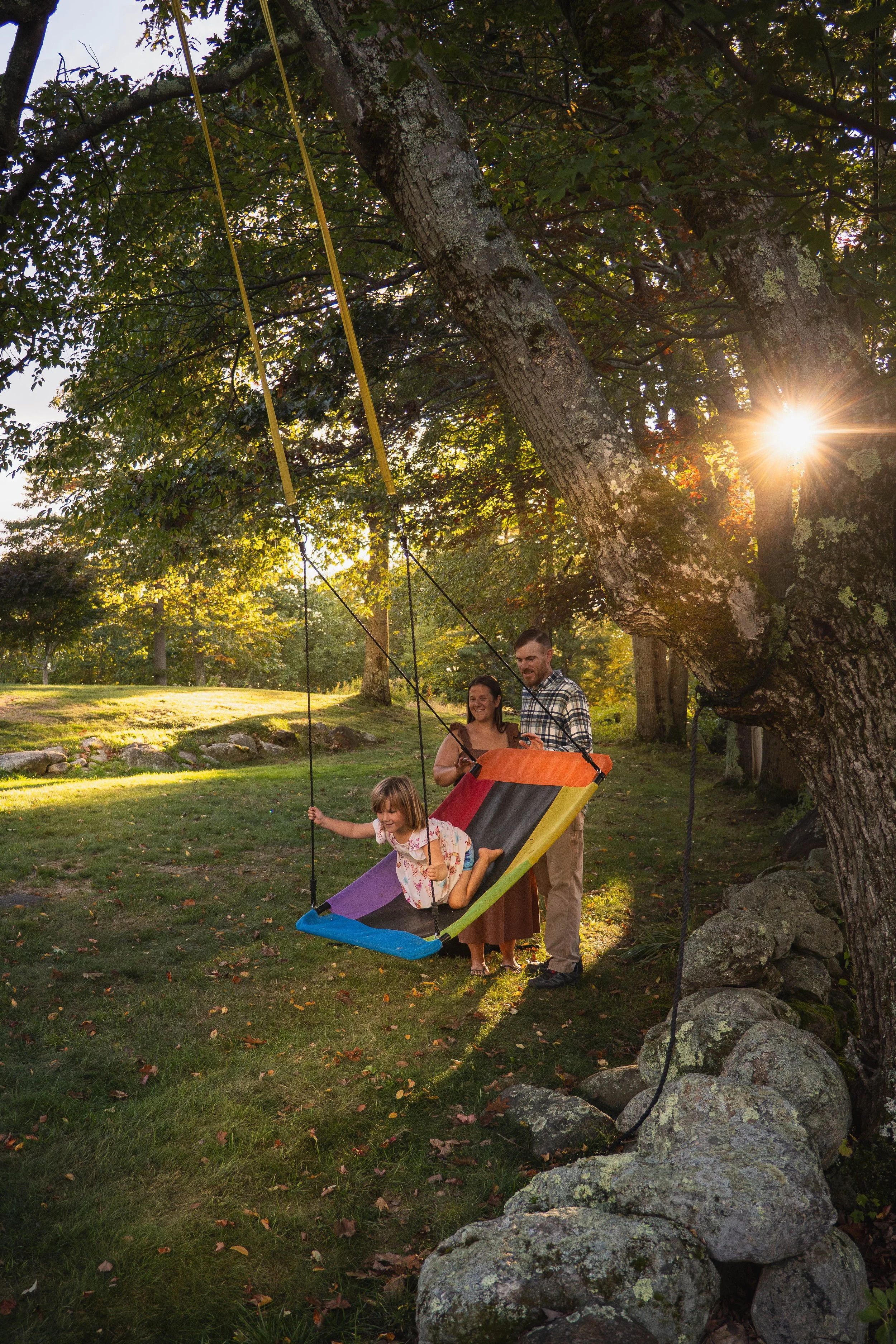 A young girl swings on a rainbow-colored tire swing attached to a large tree, with her parents standing nearby in a sunny park.
