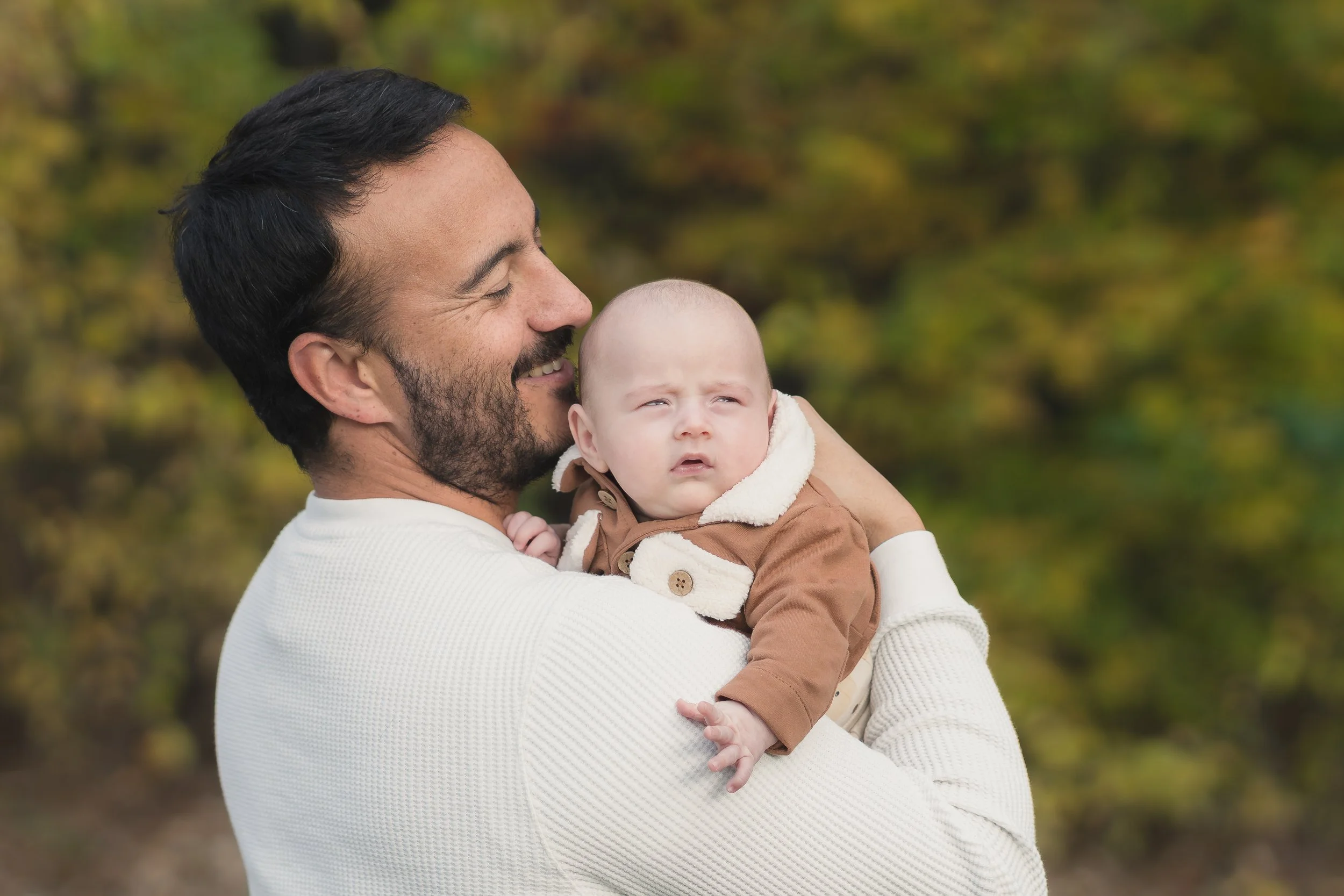 A man smiling and holding a baby outdoors with autumn foliage in the background.