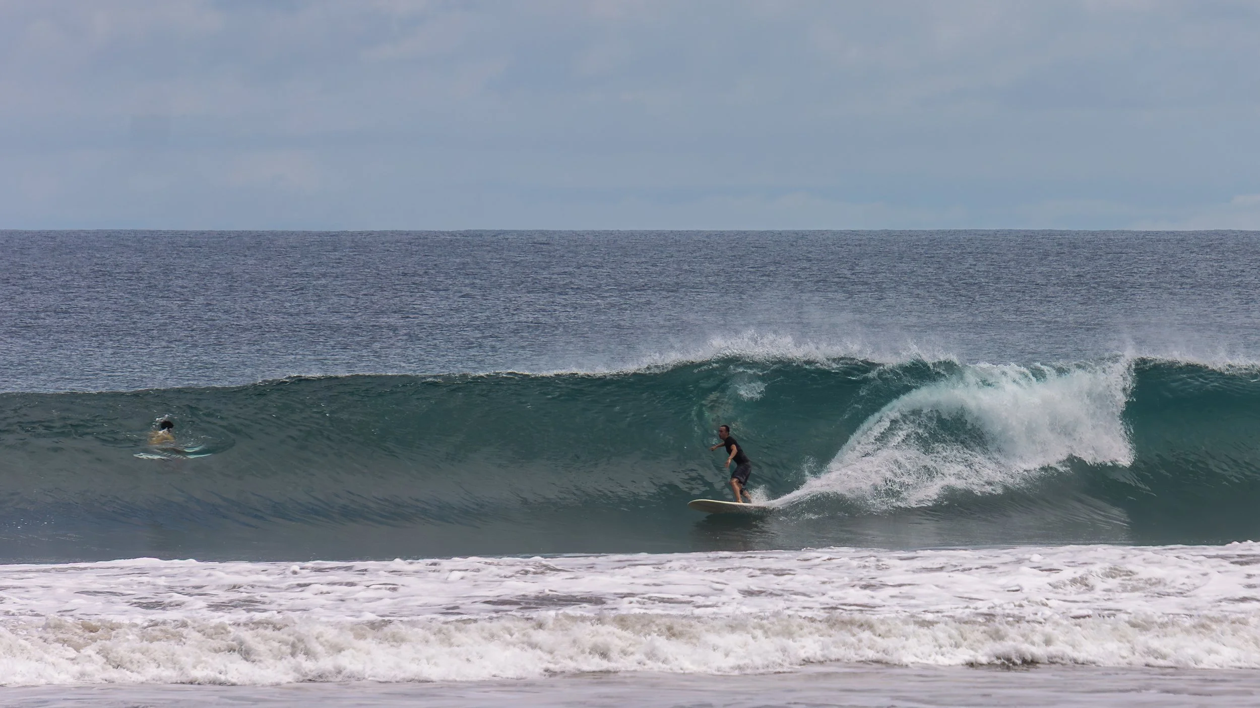 A surfer riding a wave near the beach while another person lies on a surfboard in the water.