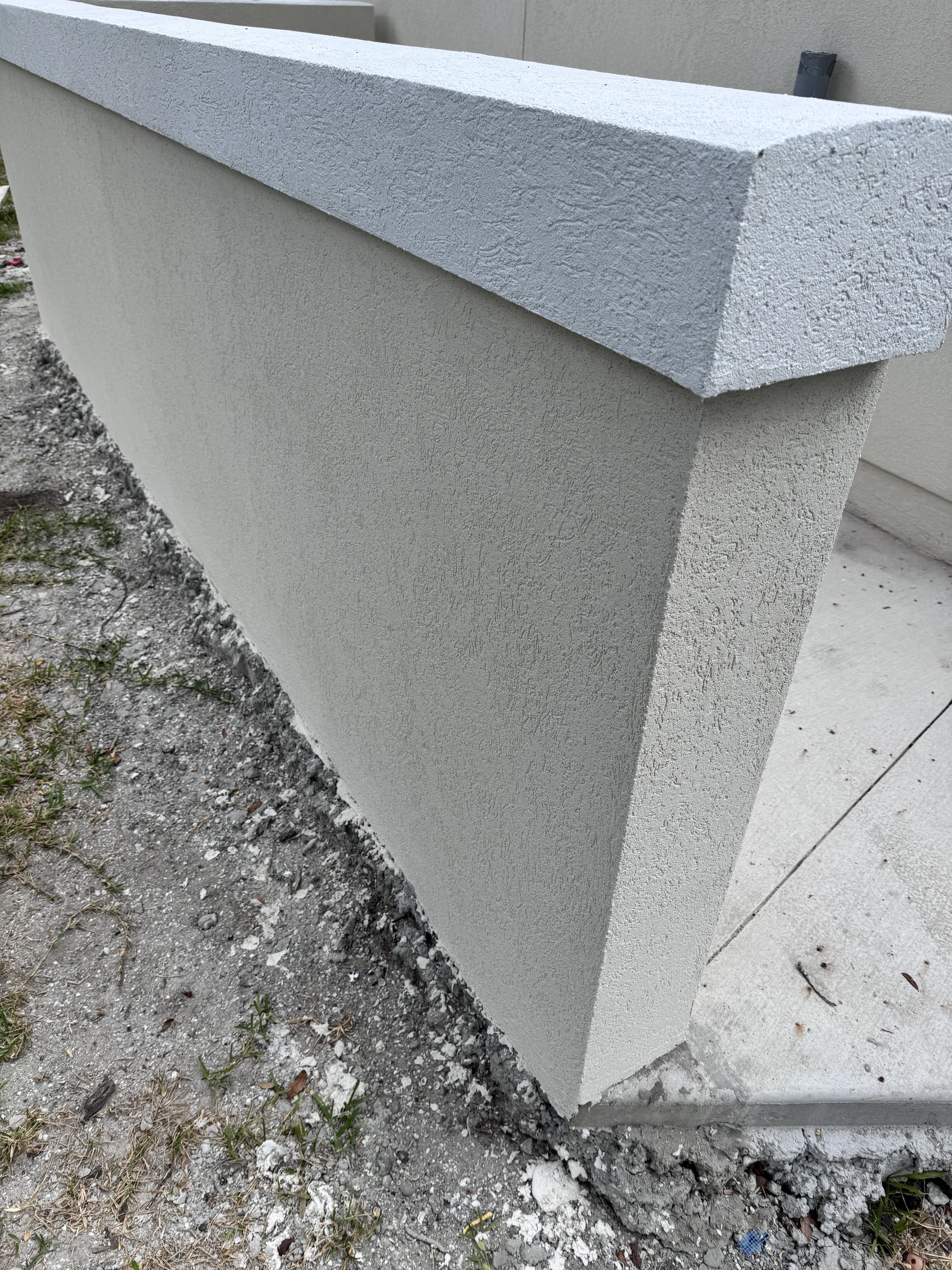 Close-up of a textured white concrete wall corner with a capped top, adjacent to dirt and grass.