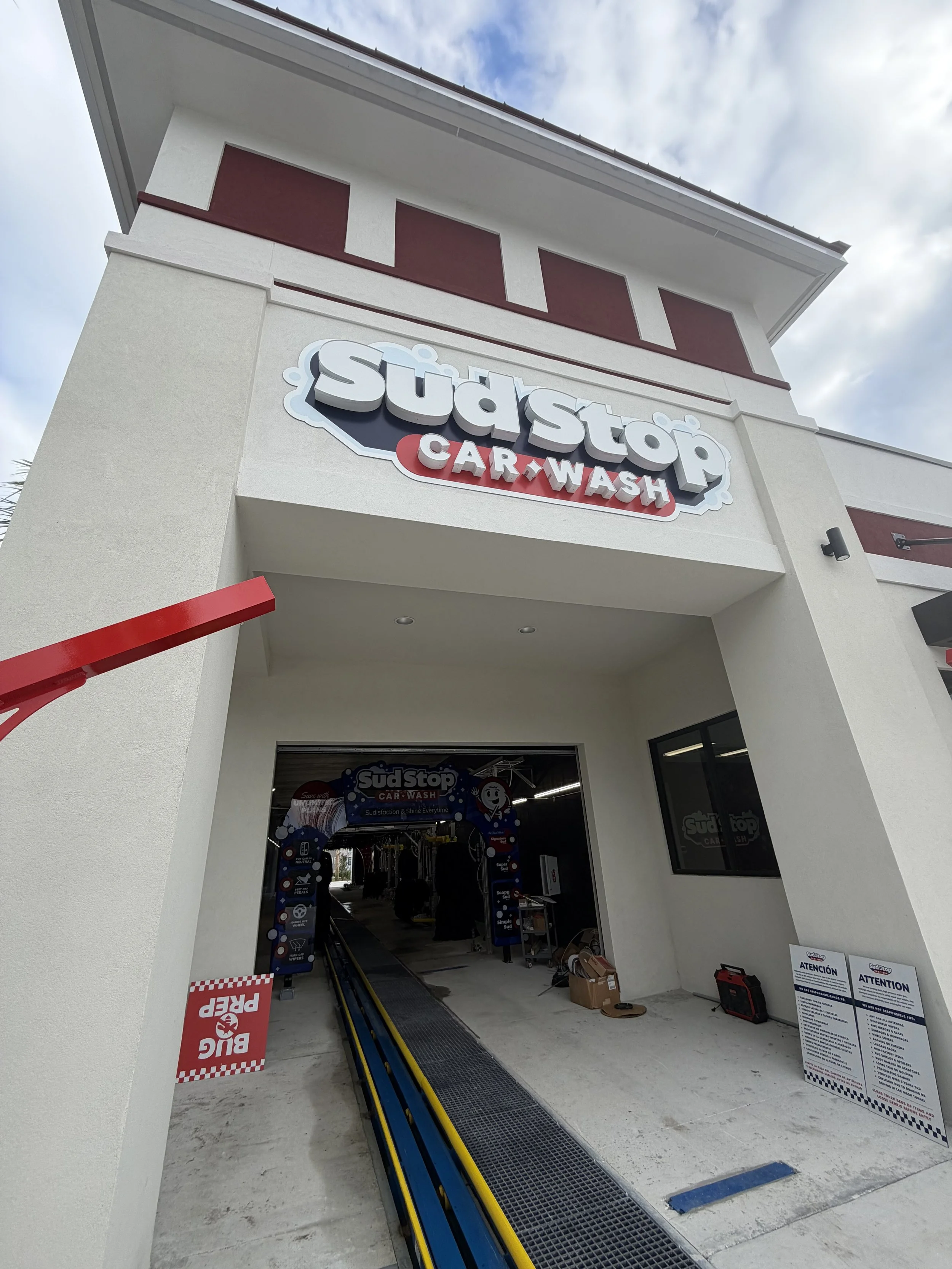 Exterior of a Sudstop car wash facility showing a ride-through wash tunnel entrance with a sign above. The building has a white facade with red accents, and a few signs and equipment are visible near the entrance.
