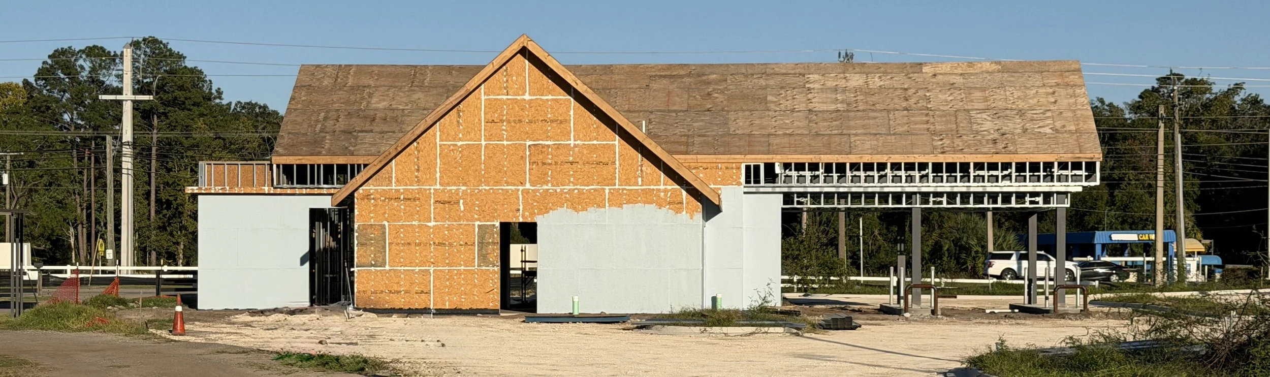 Building under construction with a new roof and partially finished walls, next to a car wash and parking lot, with trees and power lines in the background.
