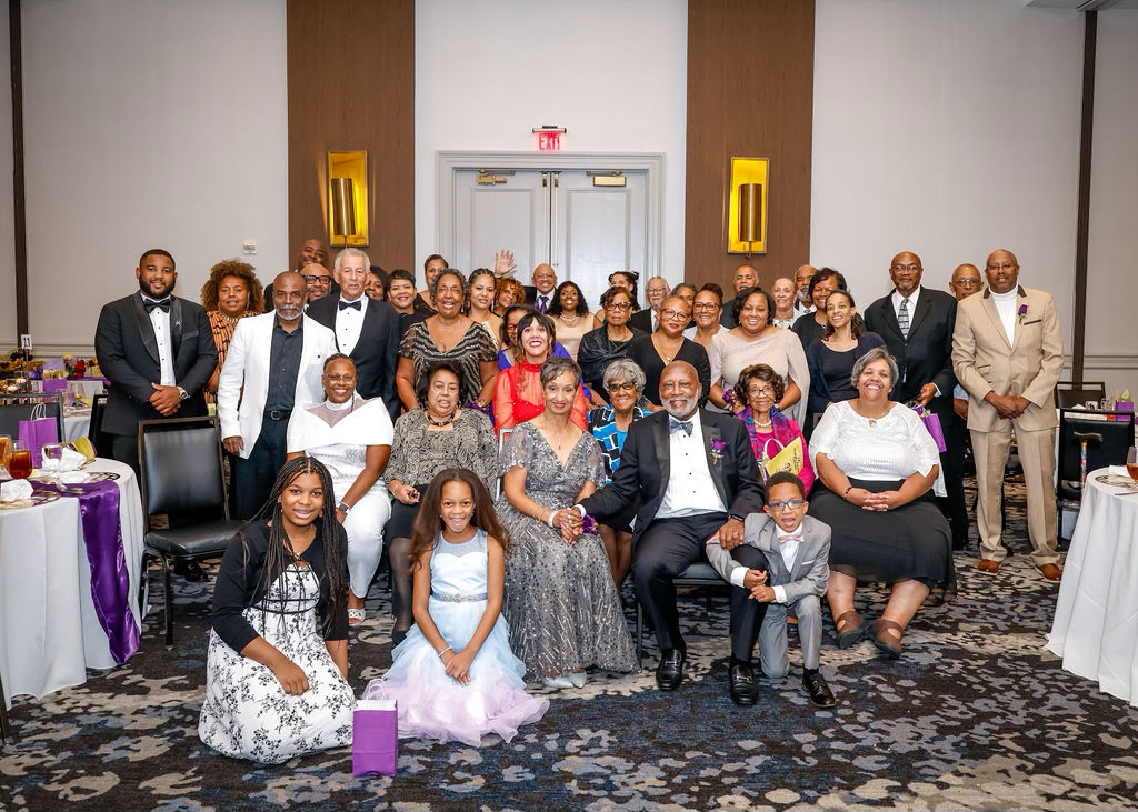 A large group of people dressed in formal attire posing for a group photo in a banquet hall at a celebration or formal event.