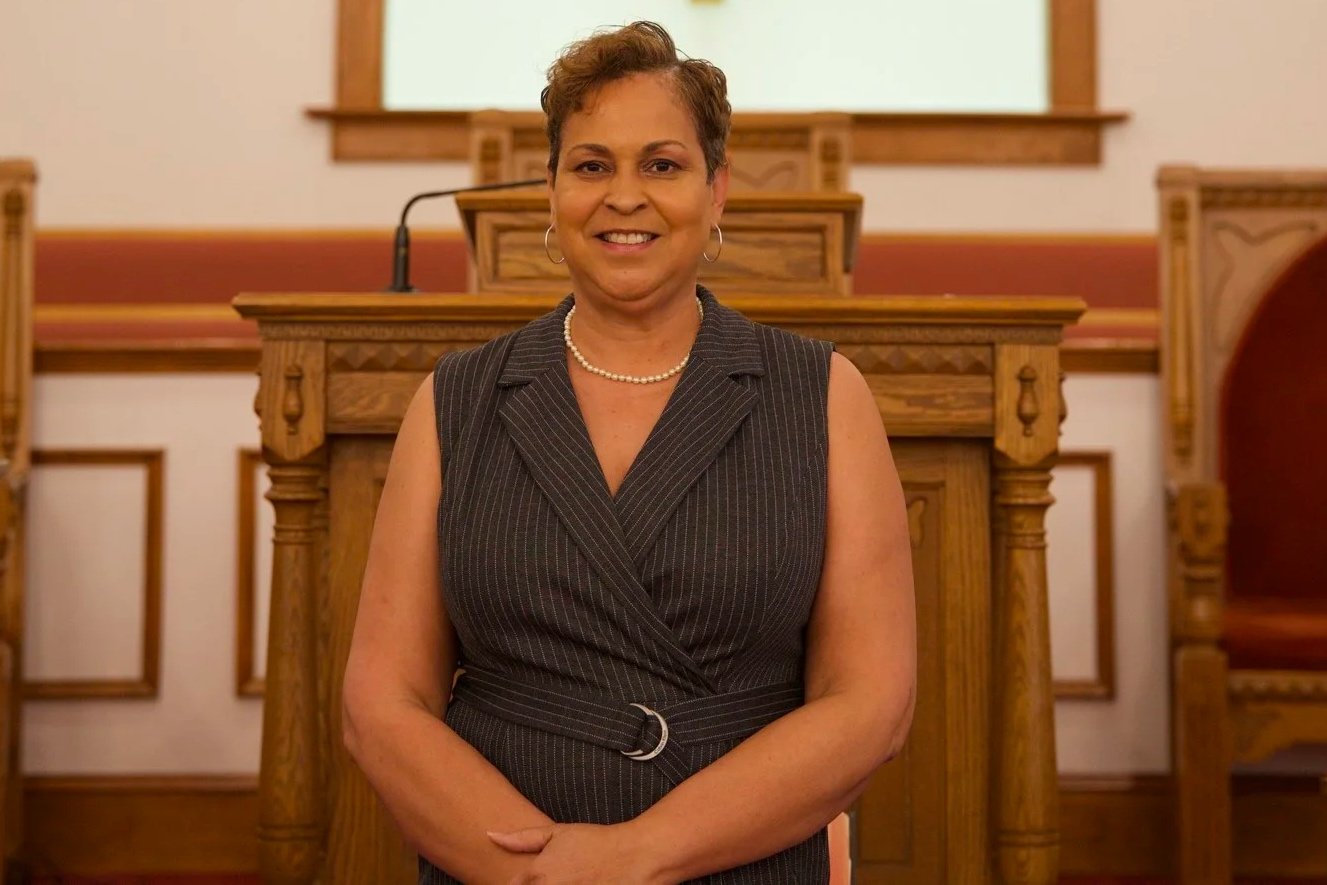 A smiling woman with short hair standing in a courtroom, dressed in a dark pinstripe sleeveless blazer, wearing pearl jewelry, with wooden courtroom furnishings in the background.