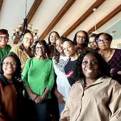 A group of women posing together indoors, smiling with a wooden-beamed ceiling and chandelier in the background.