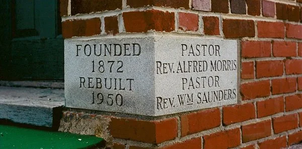 A stone corner plaque on a brick building indicating it was founded in 1872, rebuilt in 1950, with pastors Rev. Alfred Morris and Rev. Wm Saunders.