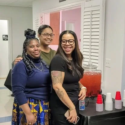 Three women standing together at a social gathering, smiling near a beverage station with red and white cups.