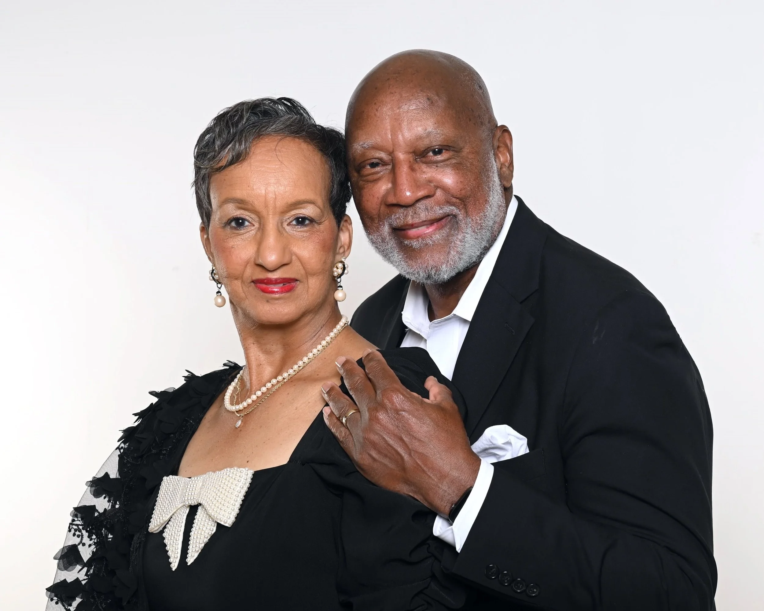 A middle-aged couple dressed in formal attire, the woman with short gray hair, pearl earrings, and a pearl necklace, and the man with a bald head and gray beard, posing against a plain white background.