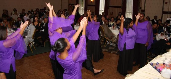 A group of women wearing purple tops and black skirts are performing a dance in a banquet hall filled with seated attendees.