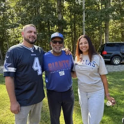 Three people standing outdoors on grass, with trees and a vehicle in the background. They are smiling and posing for the photo.