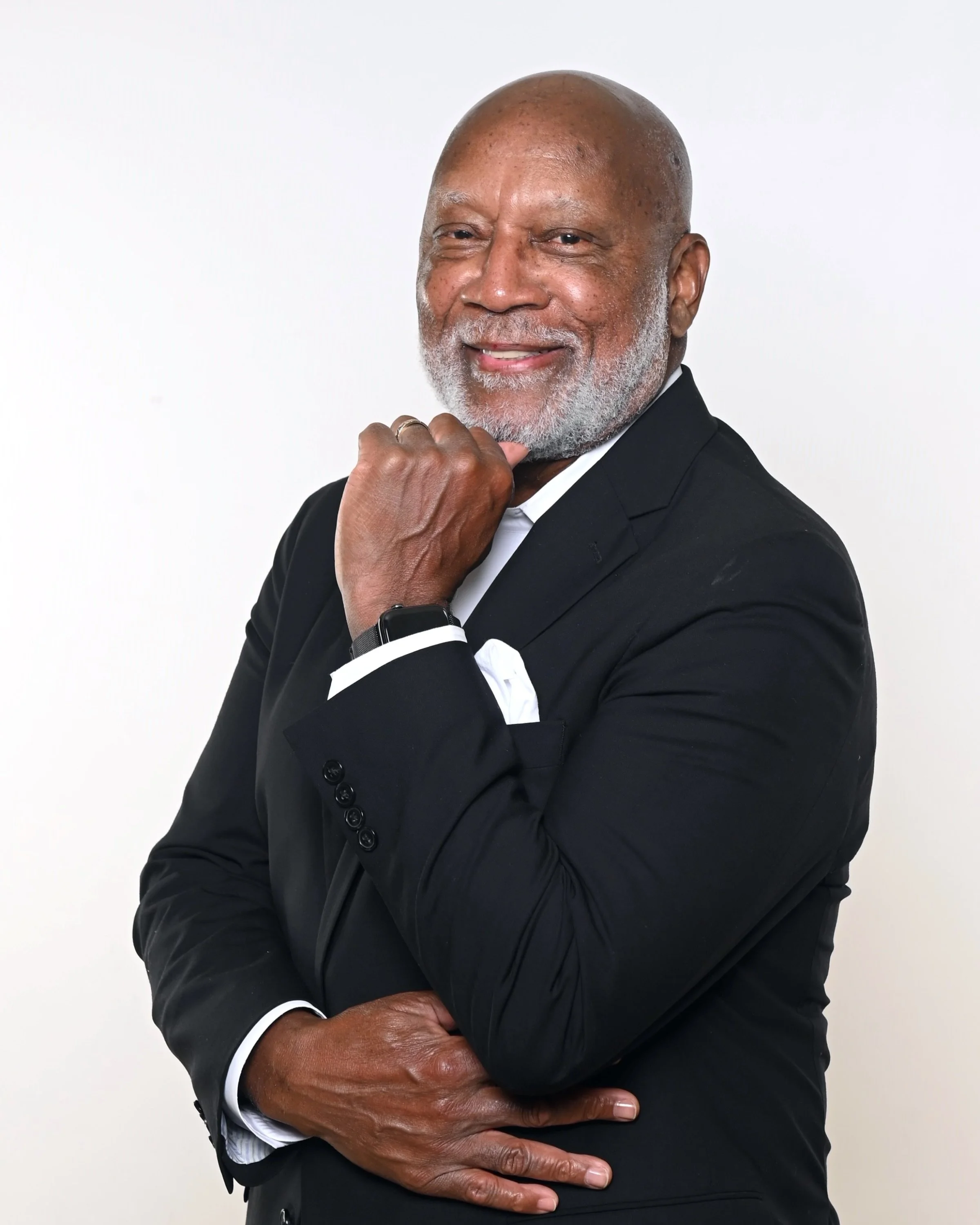 A middle-aged African American man wearing a formal black suit with a white shirt poses with his arms crossed, smiling confidently against a plain white background.