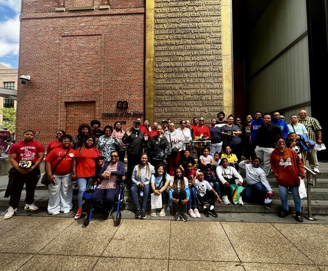Group of people posing outside the National Museum of the American Indian, with brick wall and gold plaque in the background.