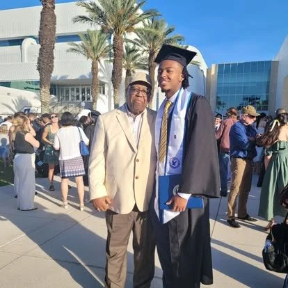 A young man in a graduation cap and gown with a blue stole, standing next to an older man in a beige blazer, at a graduation ceremony outdoors with palm trees and many people in the background.