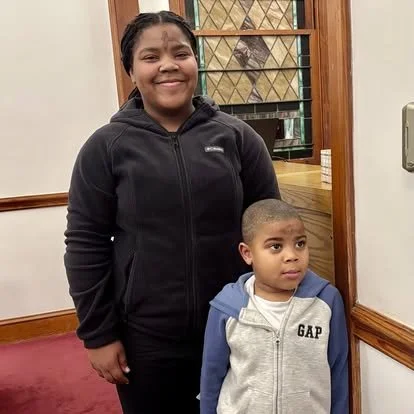 A woman standing next to a young boy inside a building, both smiling. The woman is wearing a black hoodie, and the boy is wearing a light gray and blue jacket.