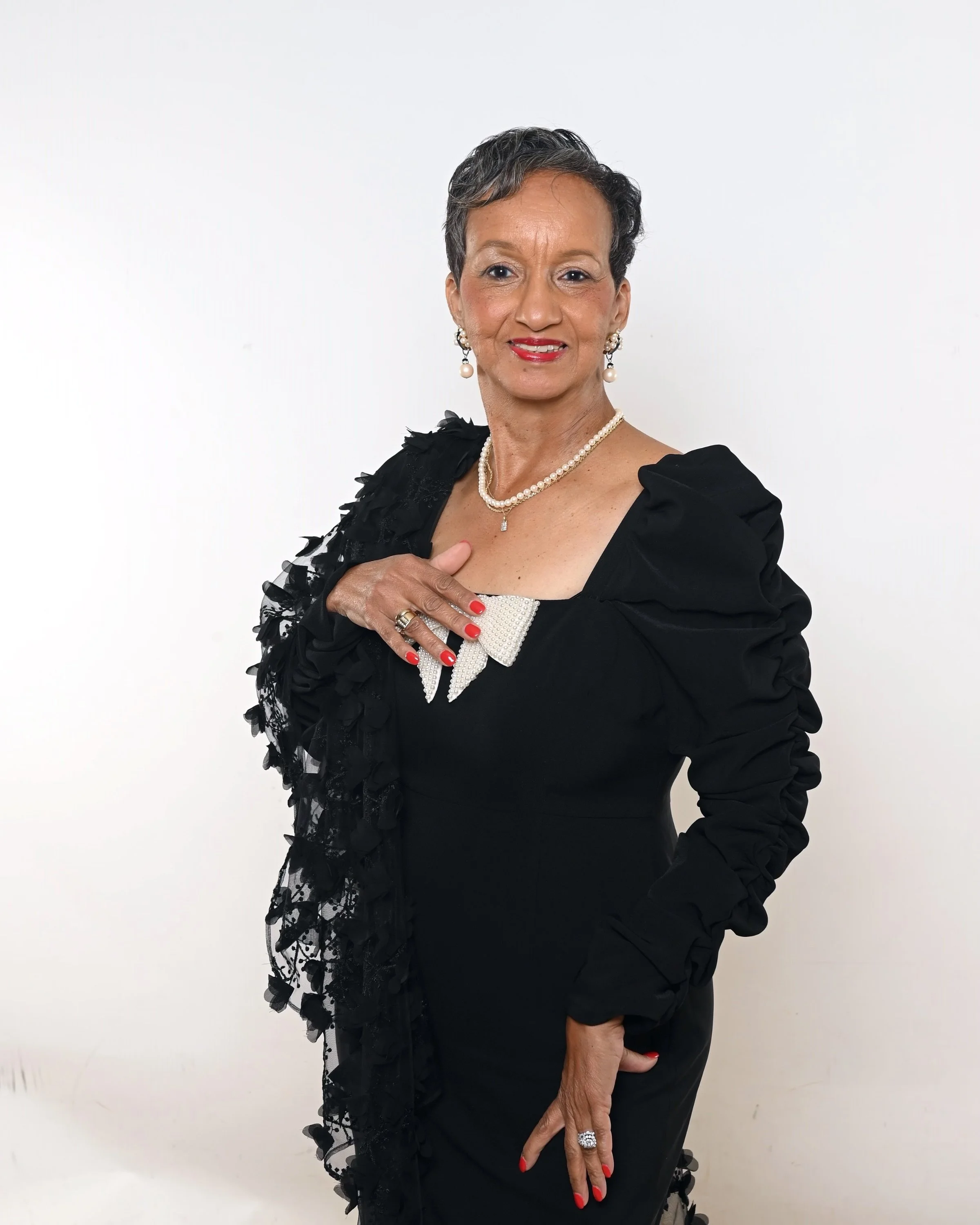A mature woman with short dark hair styled neatly, wearing a black dress with ruffled lace detailing, pearl and diamond jewelry including earrings, a necklace, and rings, standing against a plain white background. She has bright red lipstick, matching red nail polish, and is posing with one hand on her chest and the other resting on her thigh, smiling softly.