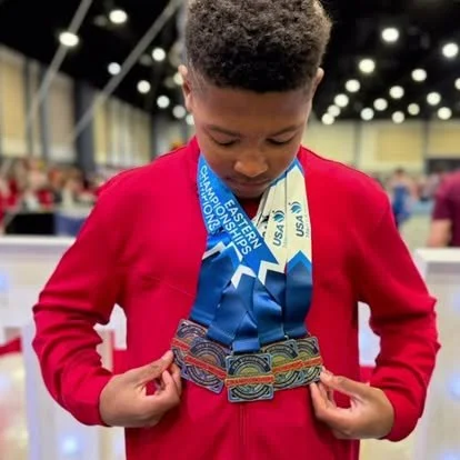 Young boy wearing a red jacket with medals around his neck at a skating rink.