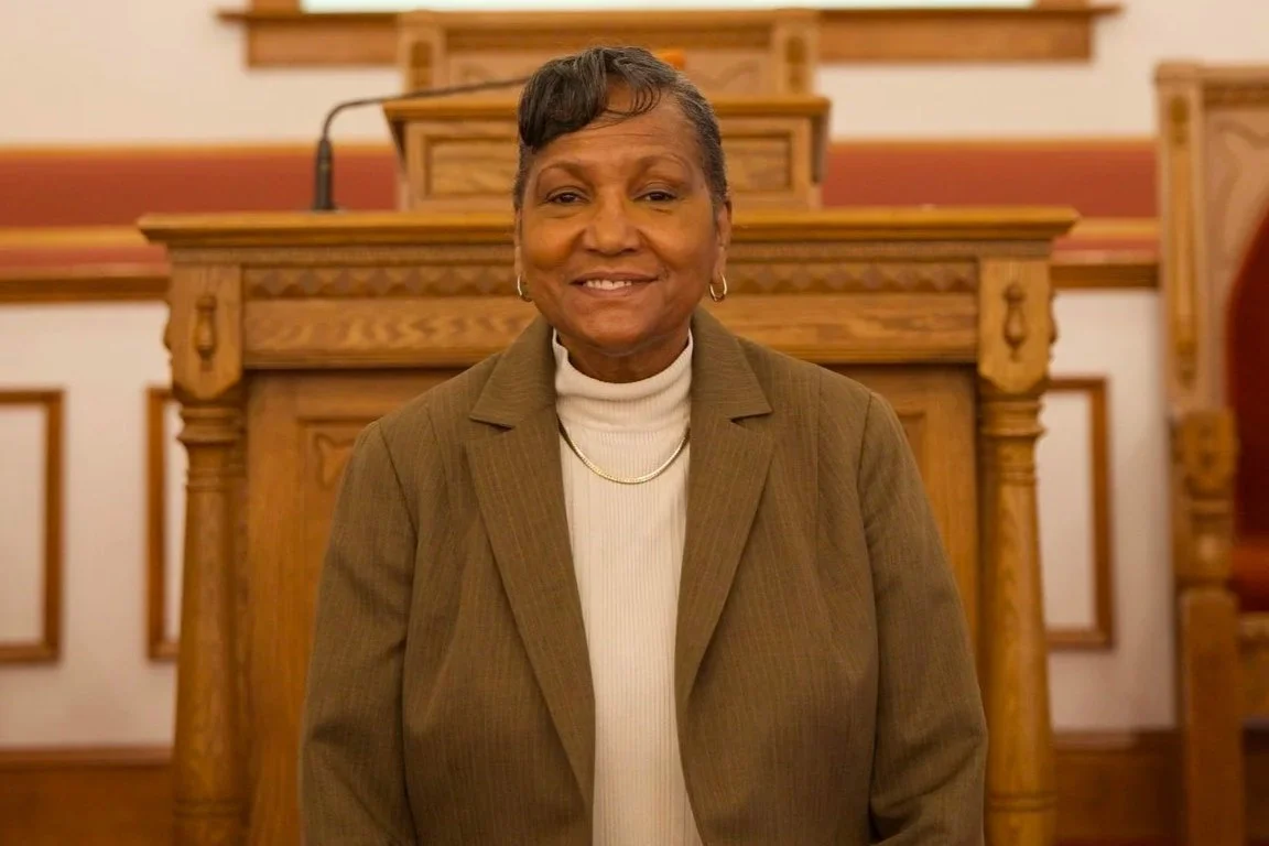 An elderly woman with short gray hair and earrings smiling, wearing a brown blazer and white turtleneck, standing in a wood-paneled courtroom or church setting.
